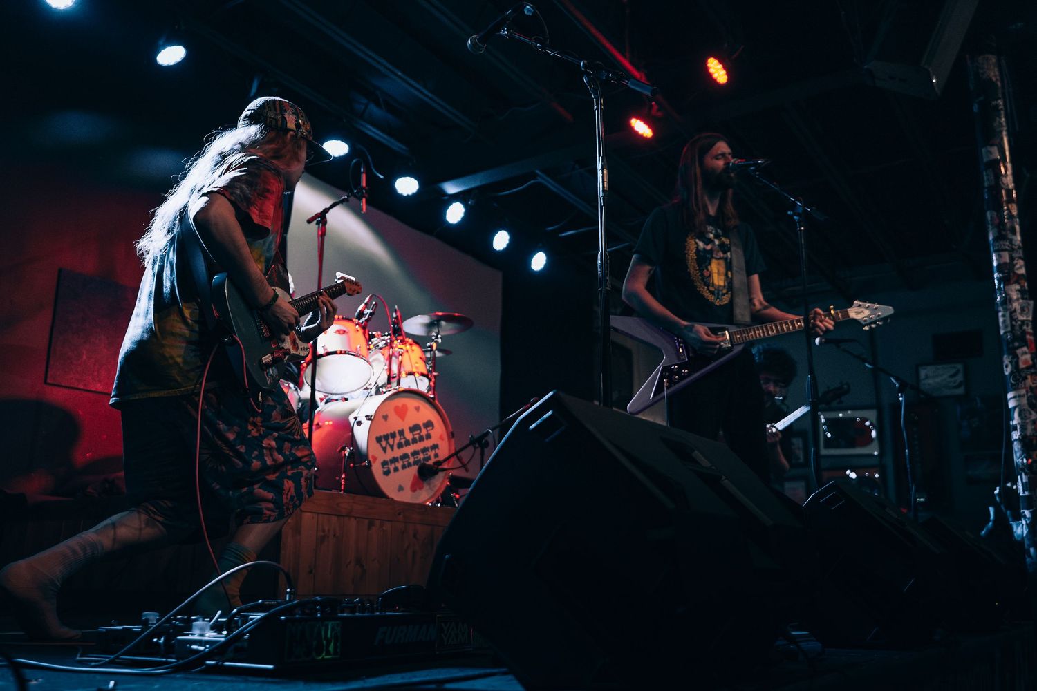 Two musicians perform on a dimly lit stage, one playing guitar while a drummer plays in the background during a live concert.