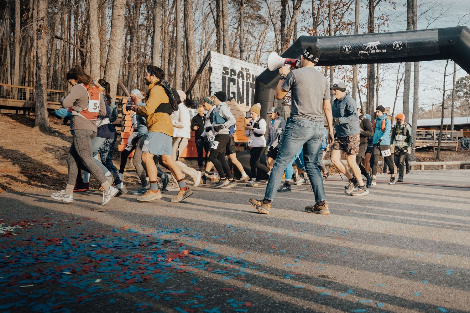 Runners crossing the start line at an outdoor race event with confetti on the ground and trees in the background.