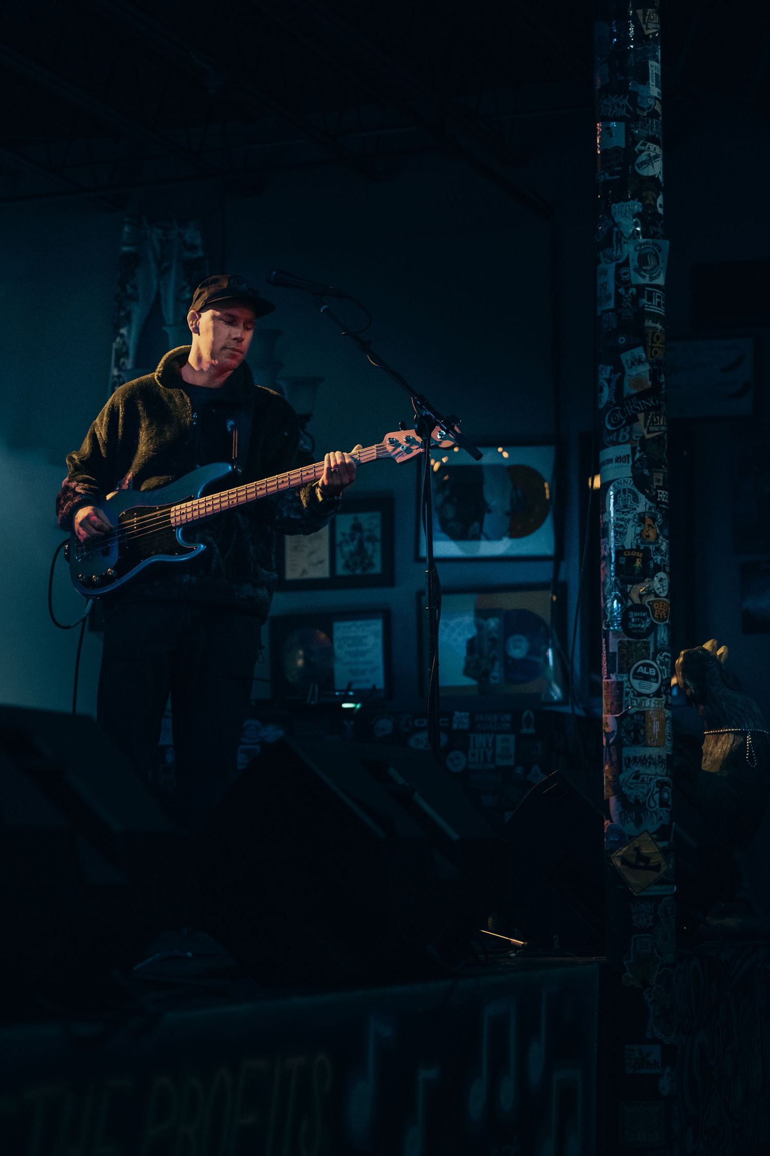 Musician playing bass guitar on dimly lit stage with sticker-covered pillar in moody blue venue lighting.