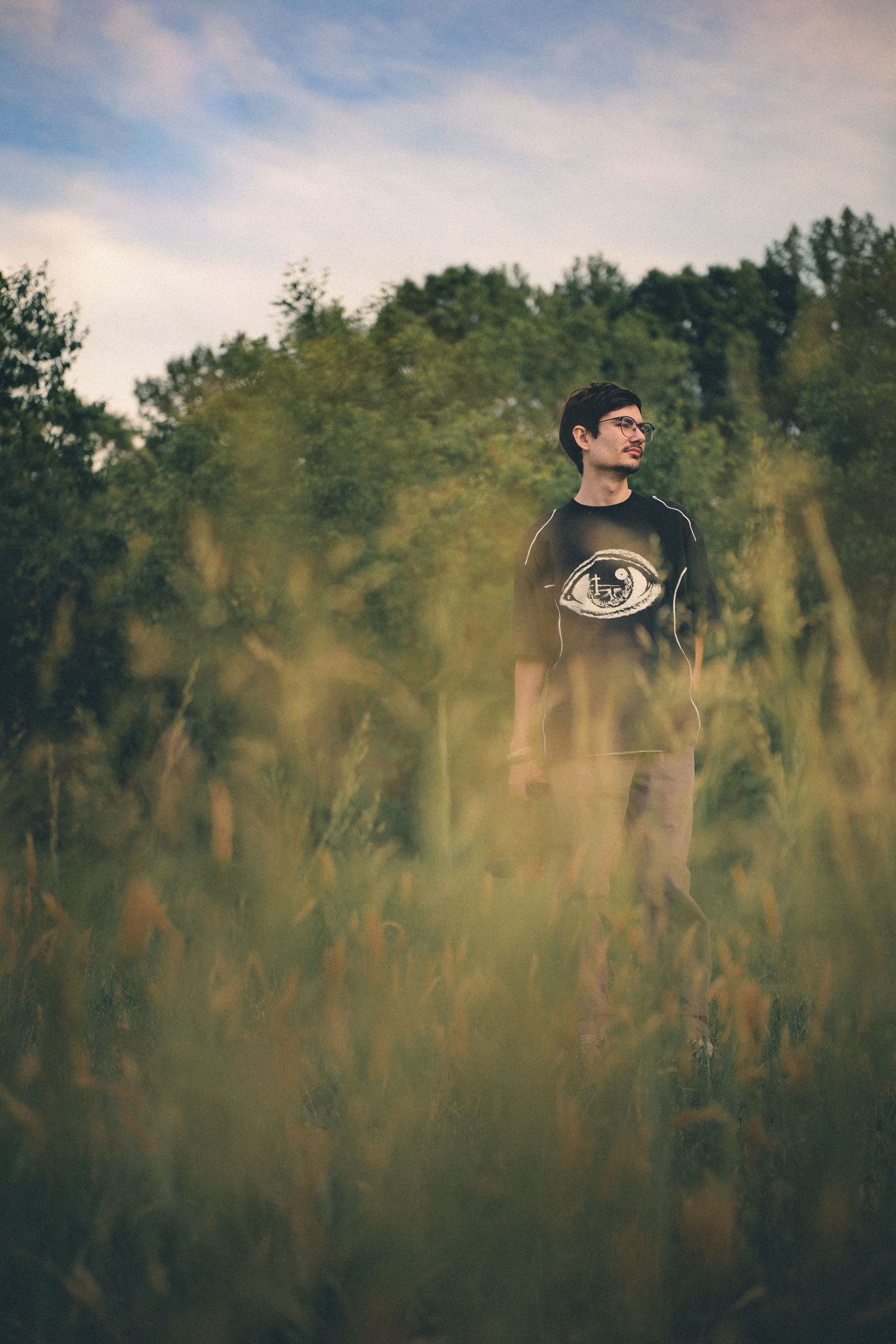 Young man in eye-print shirt standing in tall grass field with trees and cloudy sky background.