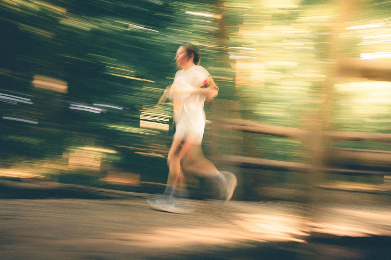 Man jogging outdoors with motion blur effect, wearing white shirt and shorts, running through a tree-lined path.