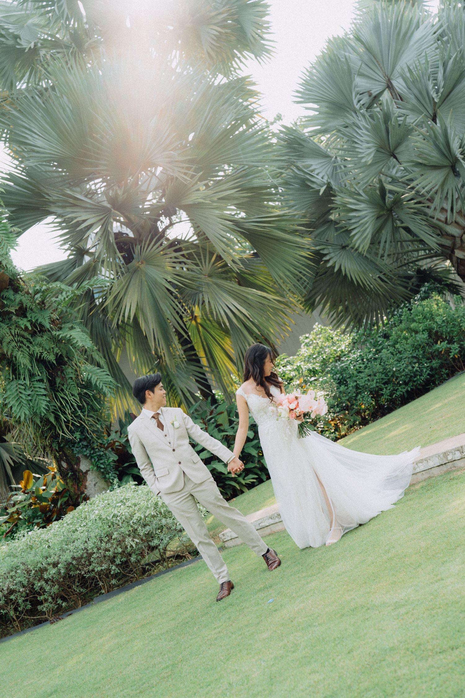 A couple in wedding attire holds hands on a lush green lawn surrounded by tropical palm trees and gardens.