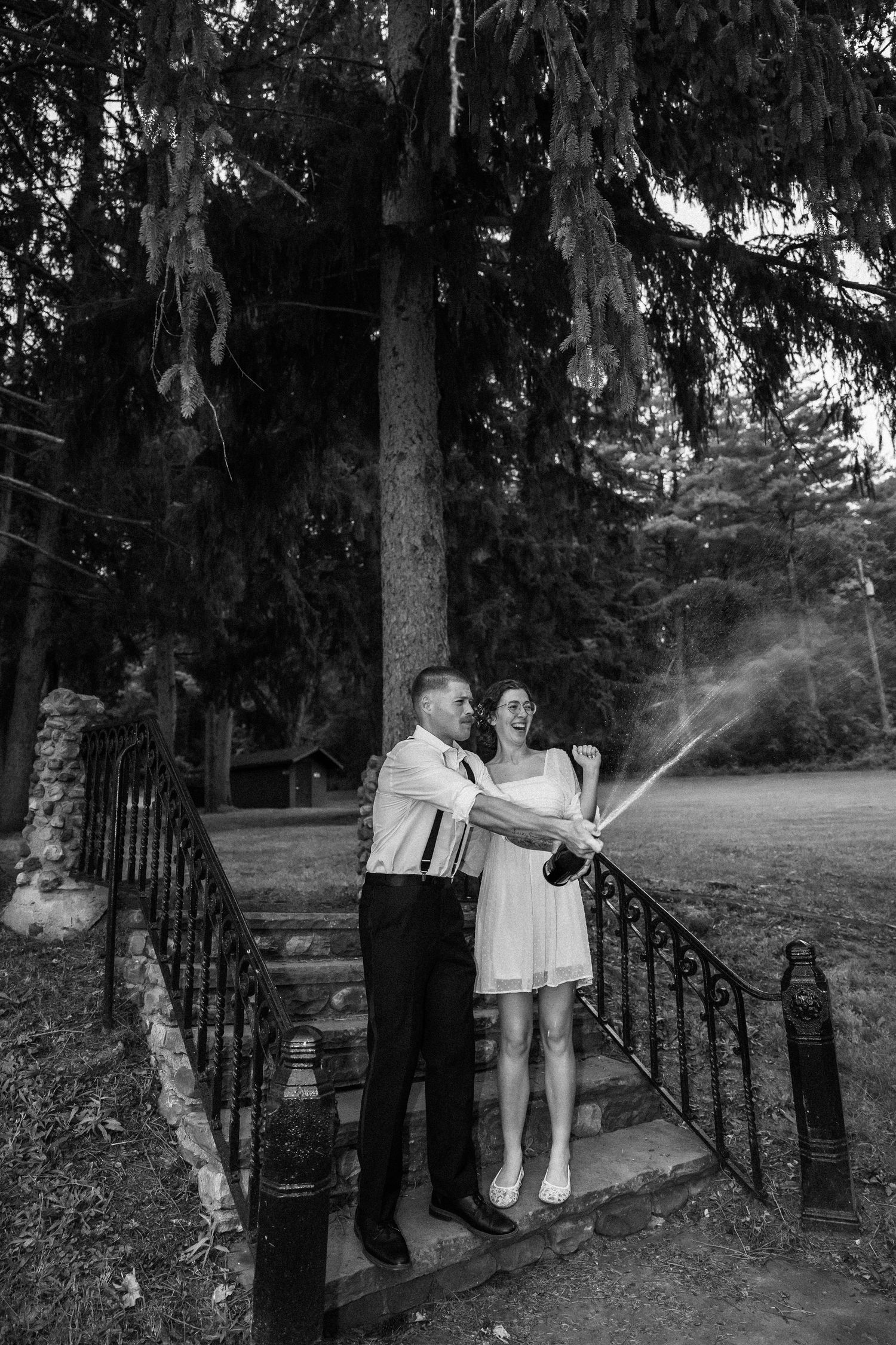 Black and white photo of a couple celebrating on outdoor stairs, popping champagne bottle near tall pine trees.