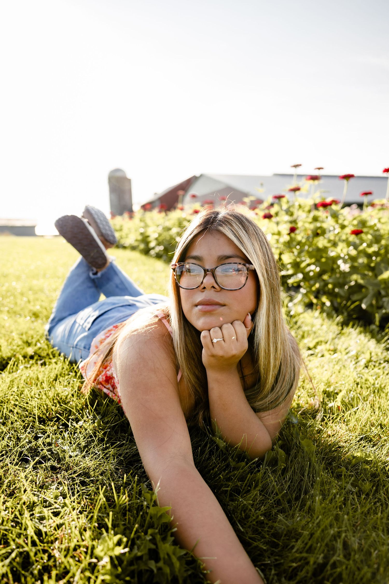 Young blonde woman with glasses lying on grass at a farm, resting chin on hand near red flowers.