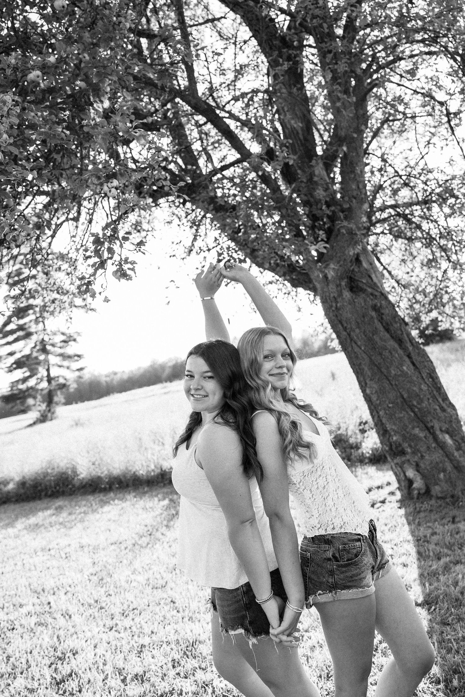 Two young women pose playfully under a large tree in a sunny field in this black and white outdoor portrait.