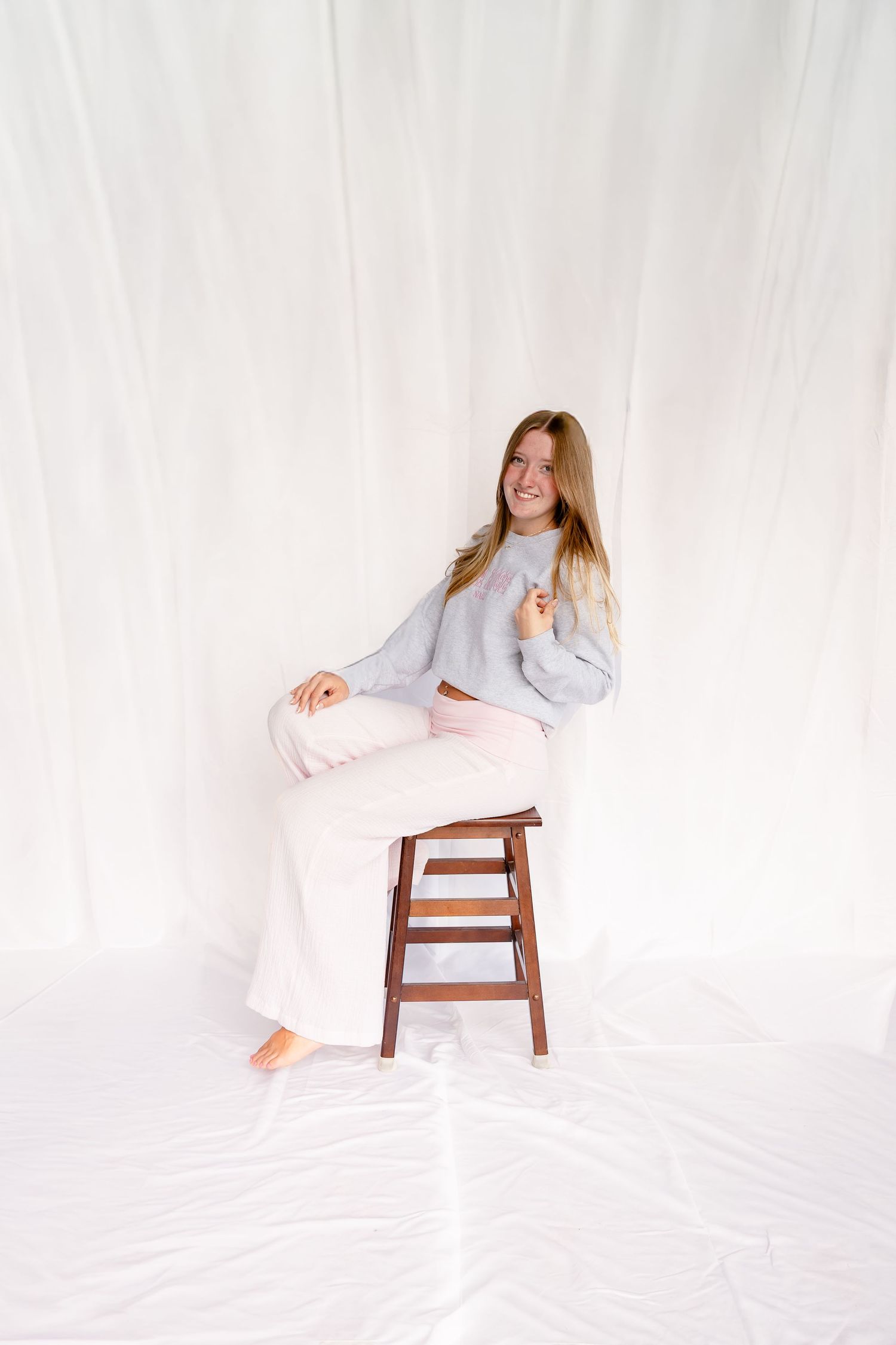 Smiling young woman in gray sweater and pink wide-leg pants sitting on wooden stool against white backdrop.