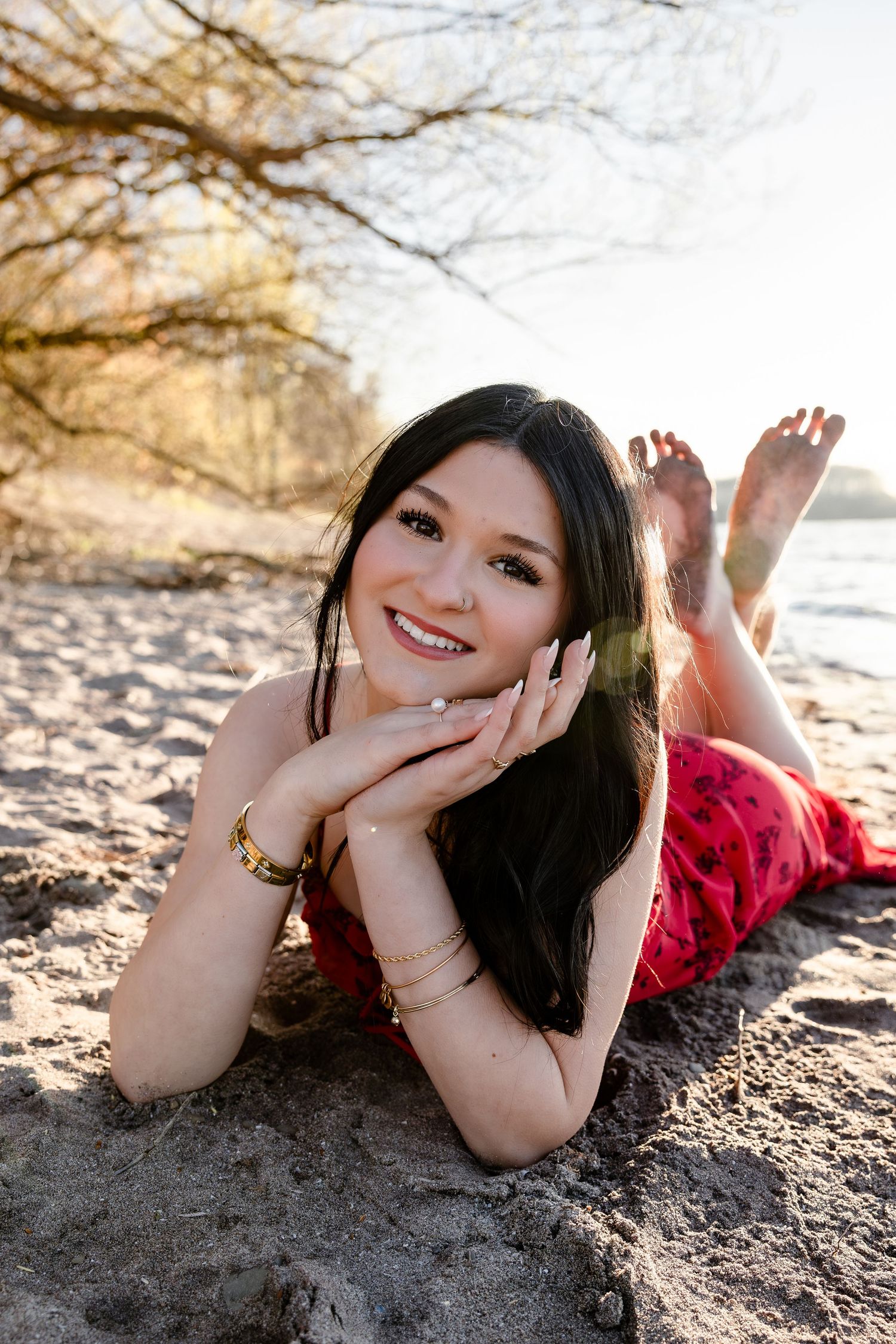 Young woman in red dress lying on sandy beach, smiling warmly with hands under chin, trees and water in background.