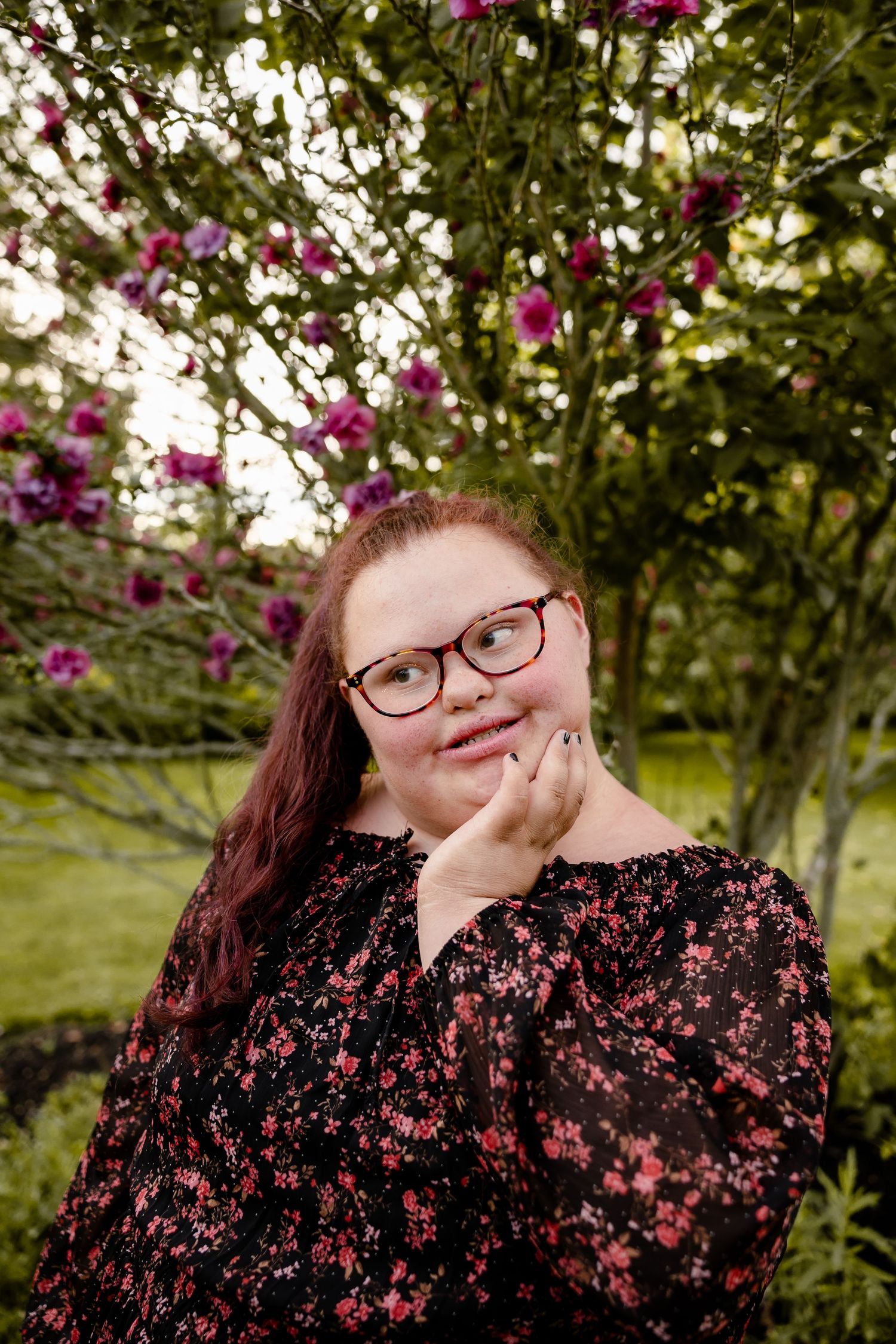 Young woman with glasses smiling in a garden with pink flowering trees, wearing a floral black dress.
