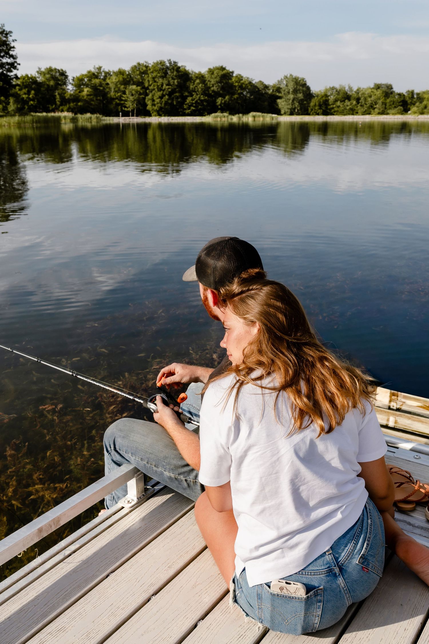 Two kids sitting on a dock fishing on a calm lake surrounded by lush green trees on a sunny day.