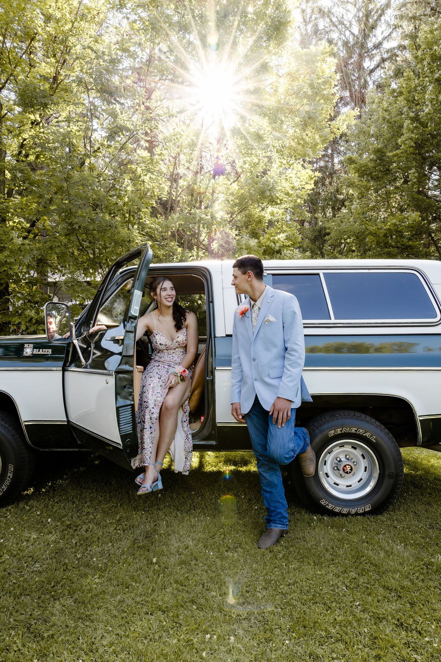 Couple in formal attire posing by a vintage white truck outdoors with sunlight streaming through green trees.
