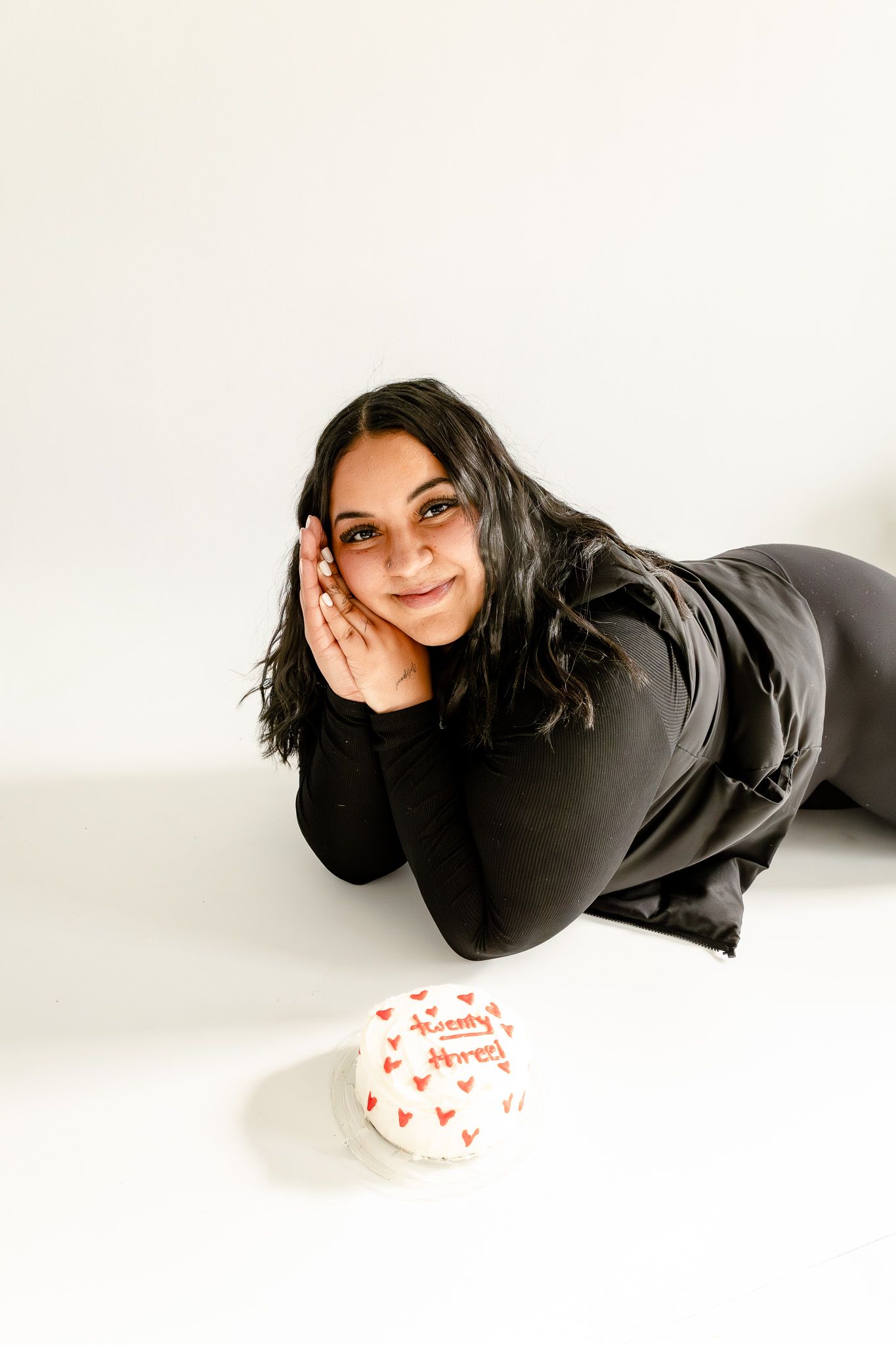 Young woman in black outfit lying on white floor smiling beside a small decorated birthday cake with red hearts.