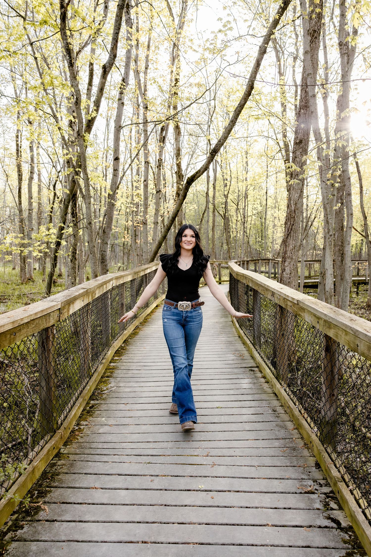 A smiling woman in jeans and black top walks along a wooden boardwalk bridge surrounded by autumn forest trees.