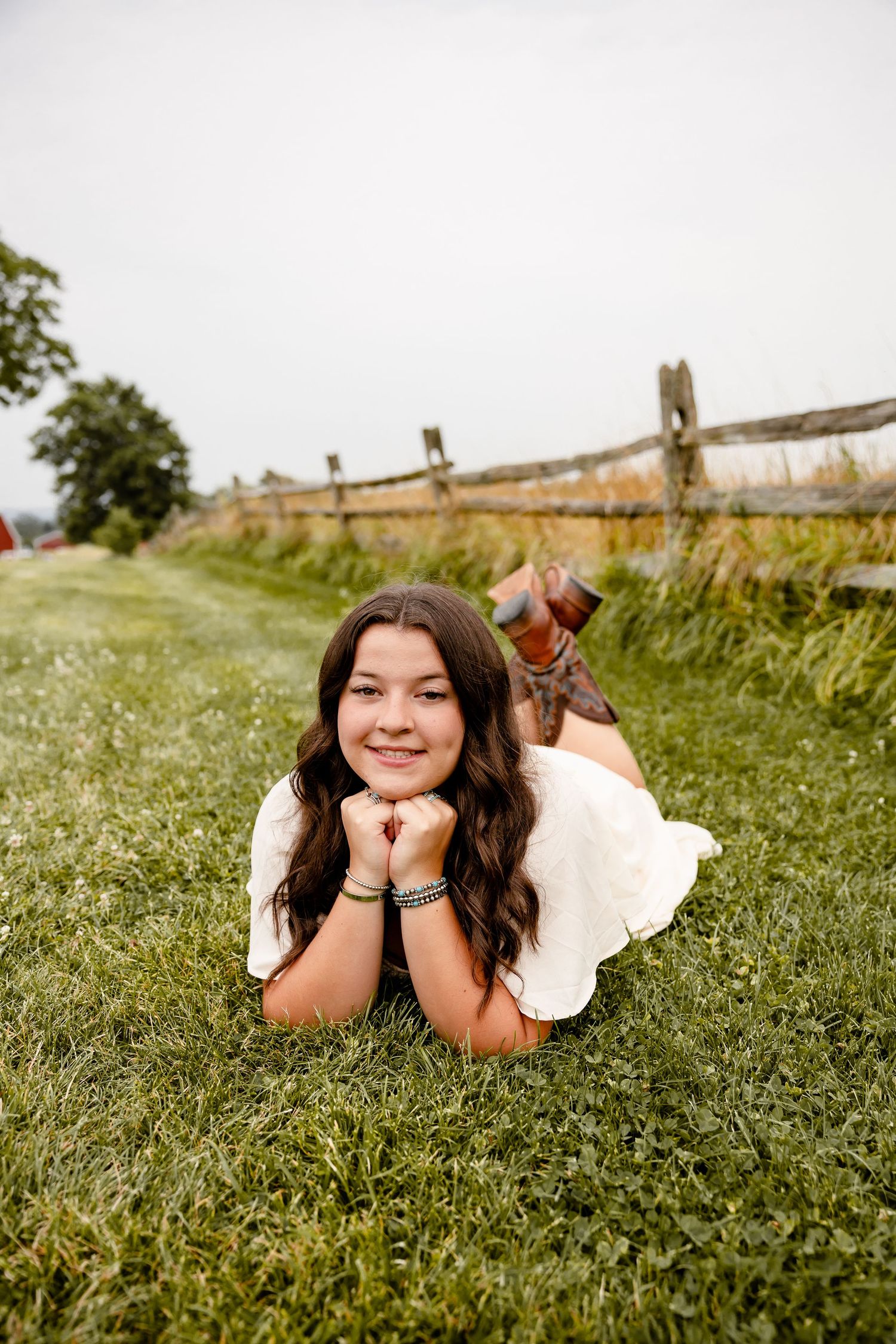 Young girl in white dress lying on grass, smiling, with rustic wooden fence and field in background.