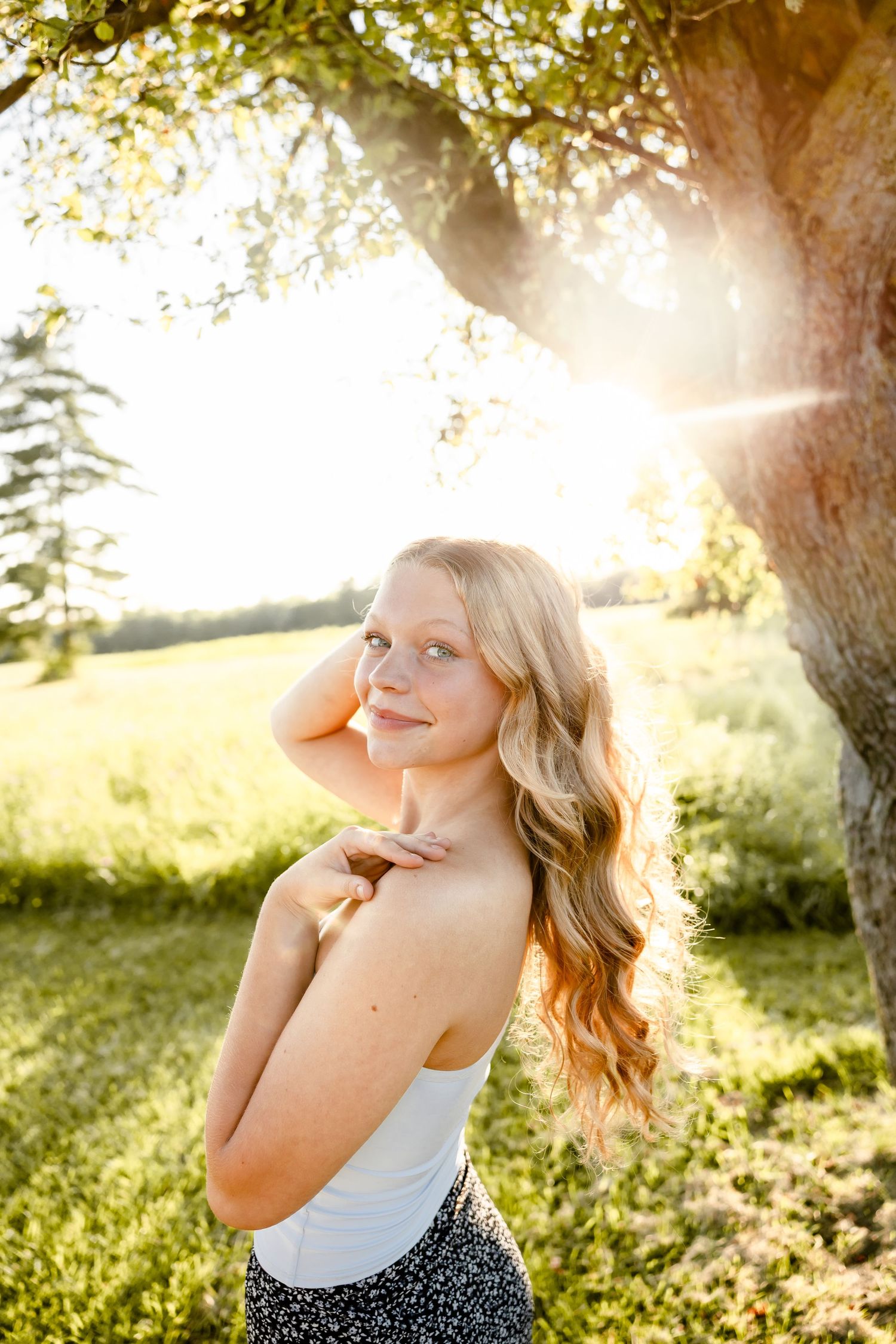 Young blonde woman in white top smiles outdoors near a sunlit tree on a bright summer day.