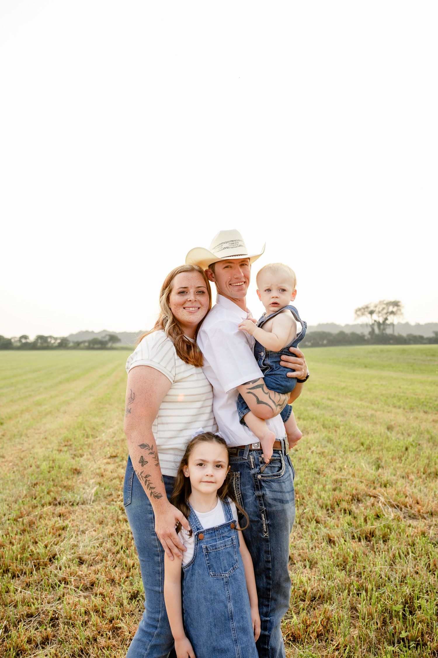 Family of four posing in a green field, parents with two young children, wearing casual denim outfits on a sunny day.