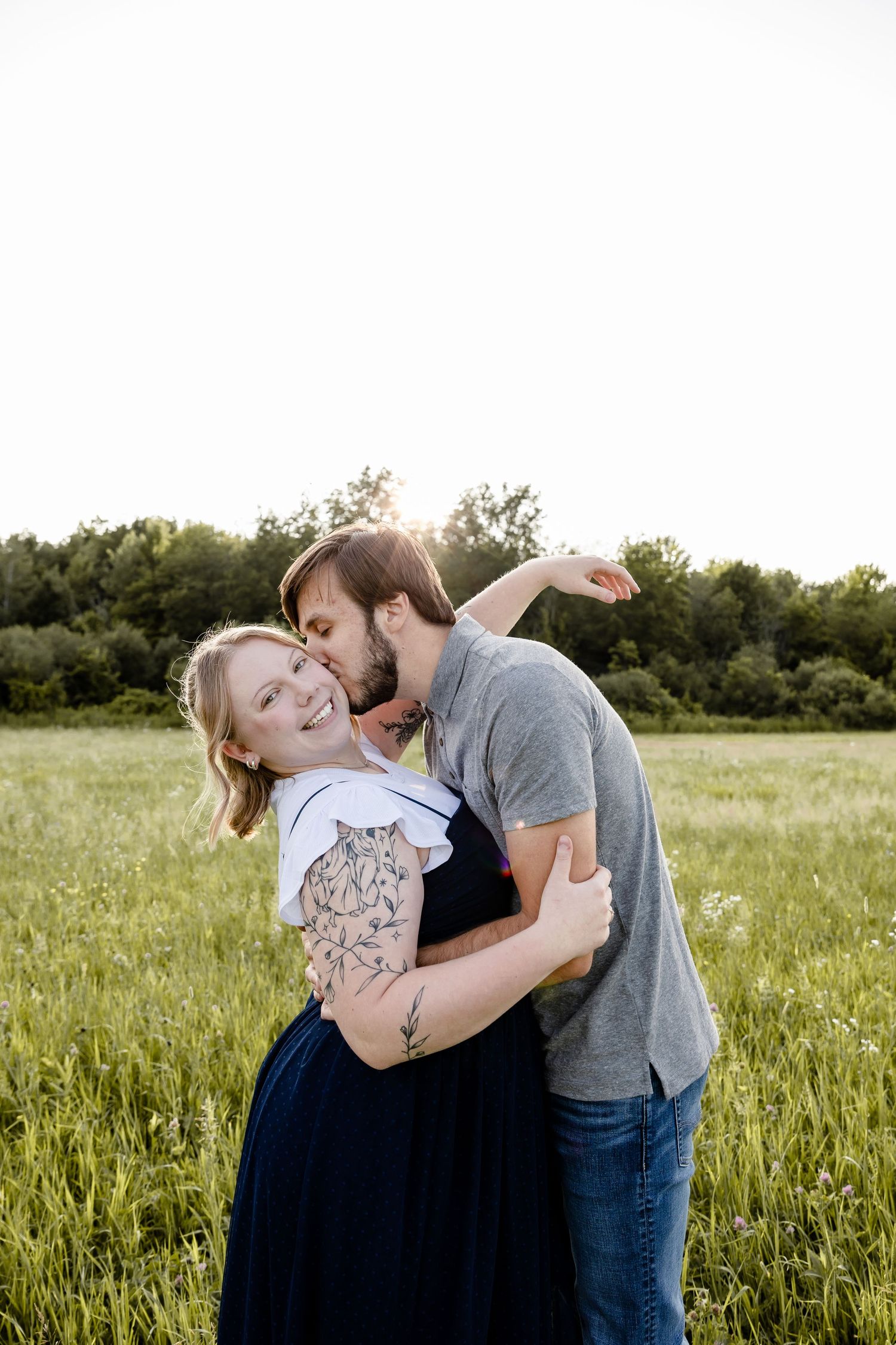 A couple embraces in a green field, man kissing woman's cheek as she smiles, with trees in the background.