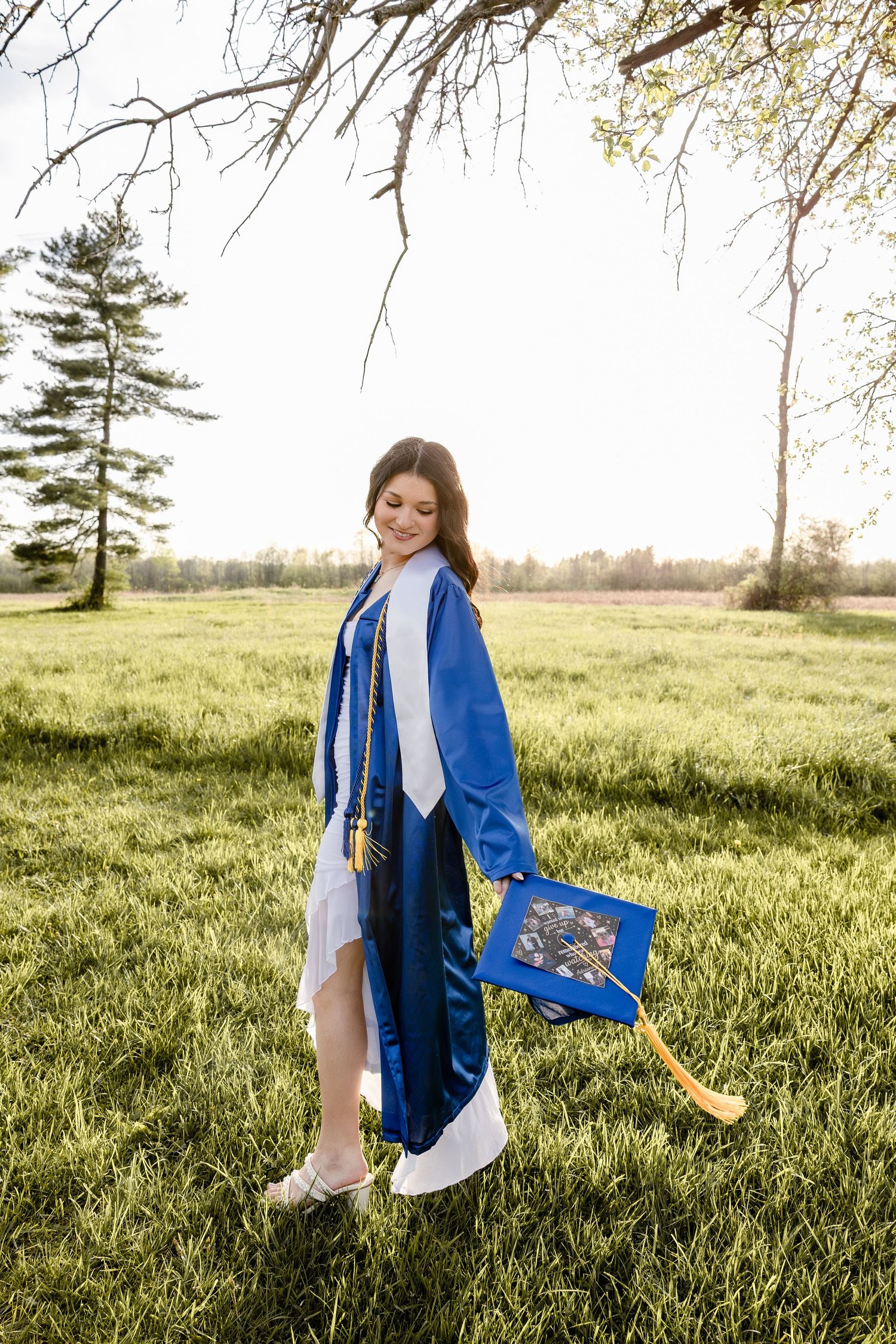 Young woman in blue graduation gown and cap smiling in a green field on a sunny spring day.