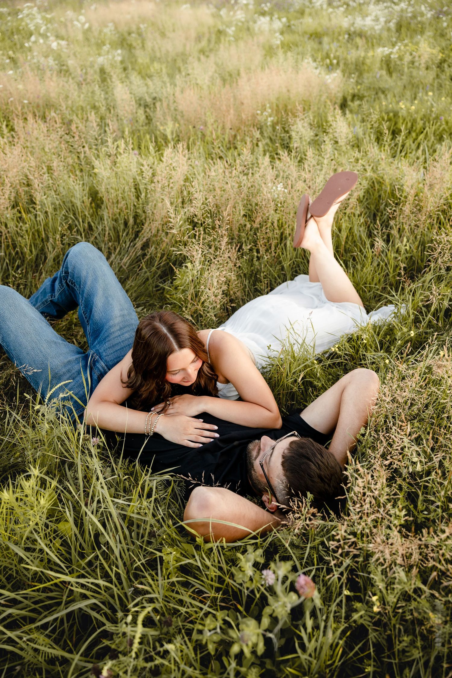Couple laying in tall green grass field, woman resting on mans chest, romantic outdoor photo session.