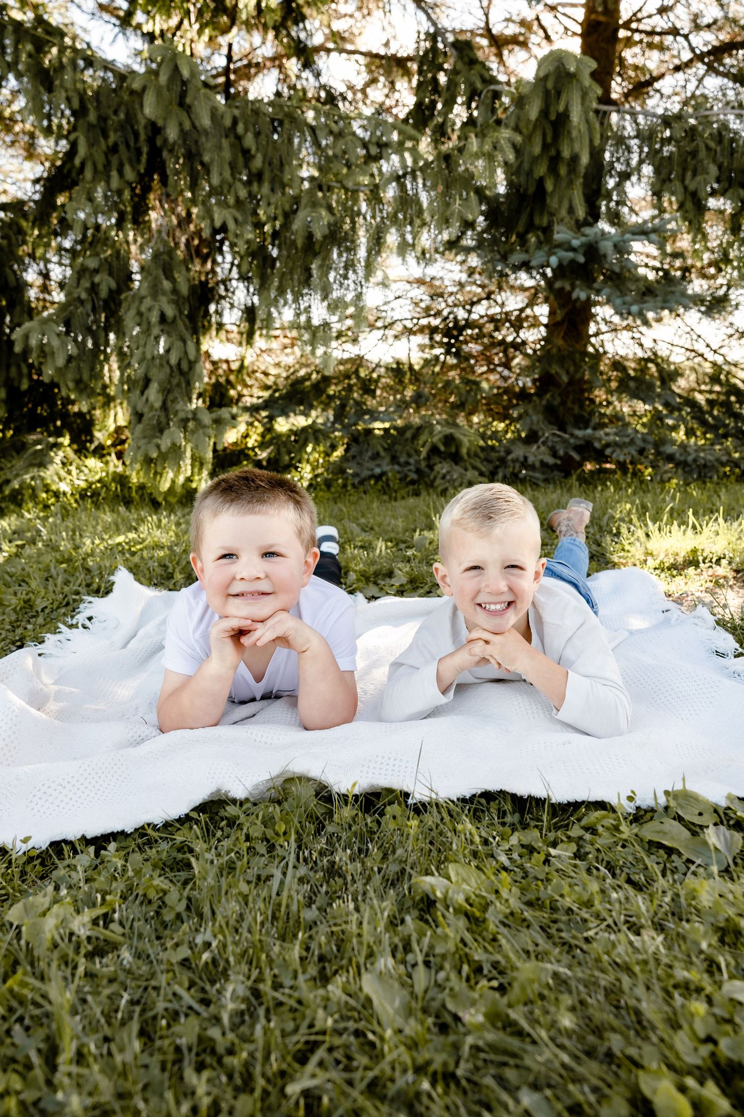 Two smiling boys lying on a white blanket in a sunny green park, posing with chins resting on hands.