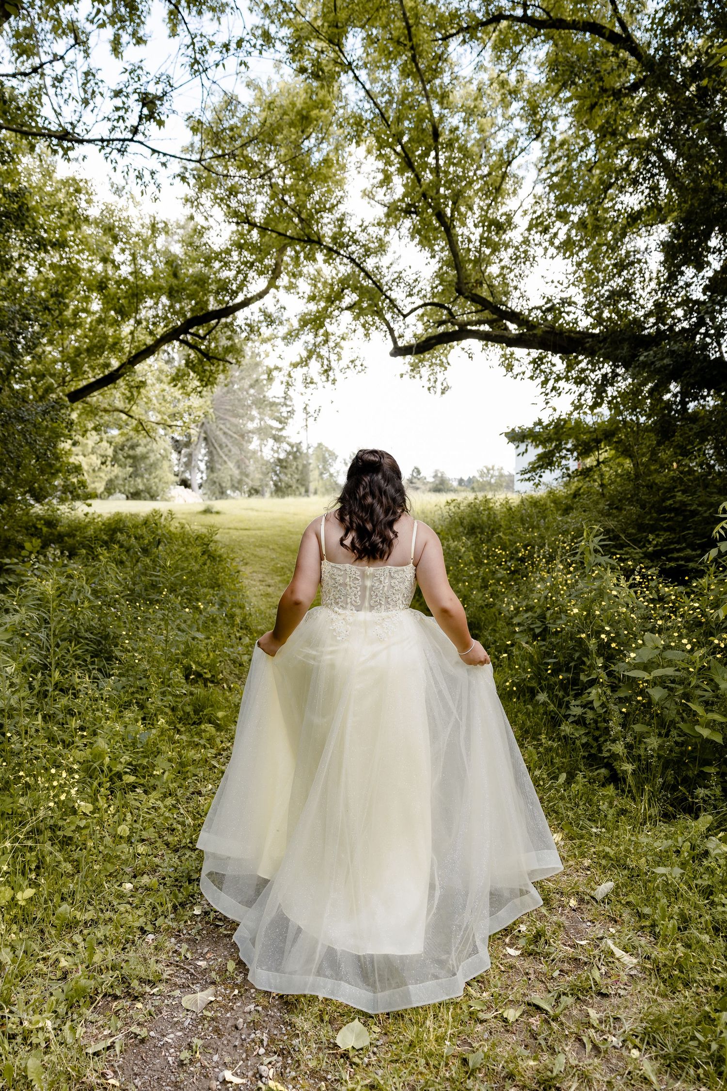 Bride in white gown walks away on garden path, holding dress, surrounded by lush green trees and foliage.