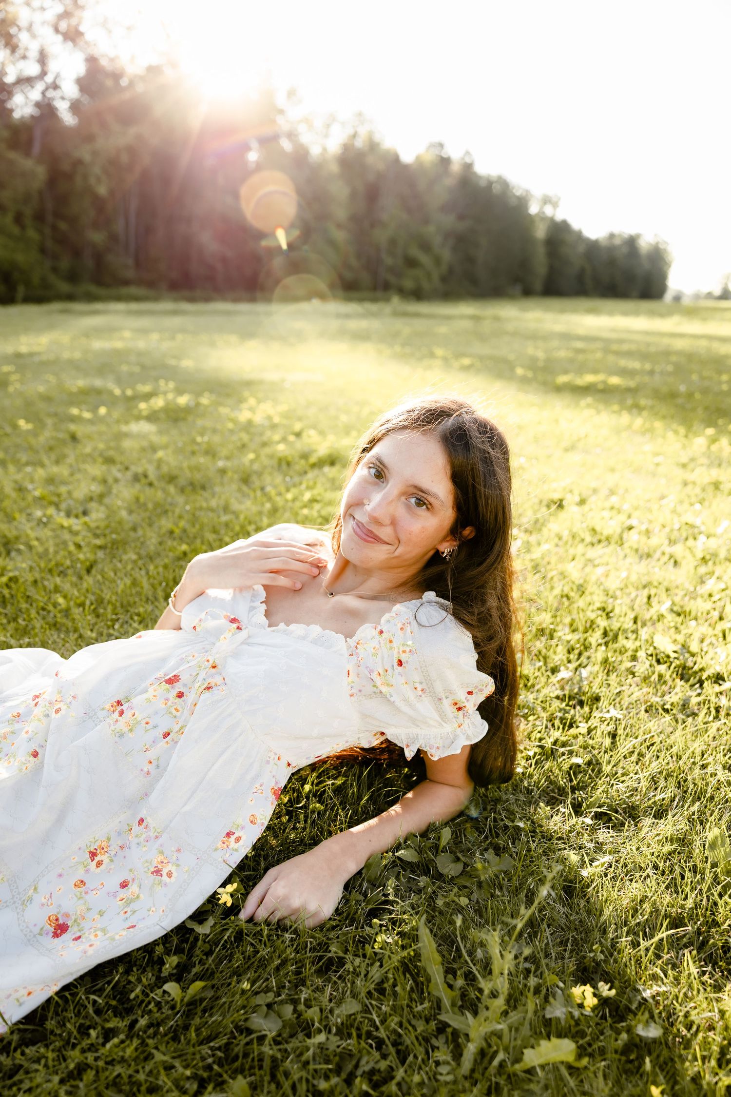 Young woman in floral white dress lying on green grass in golden sunlight with trees in background.