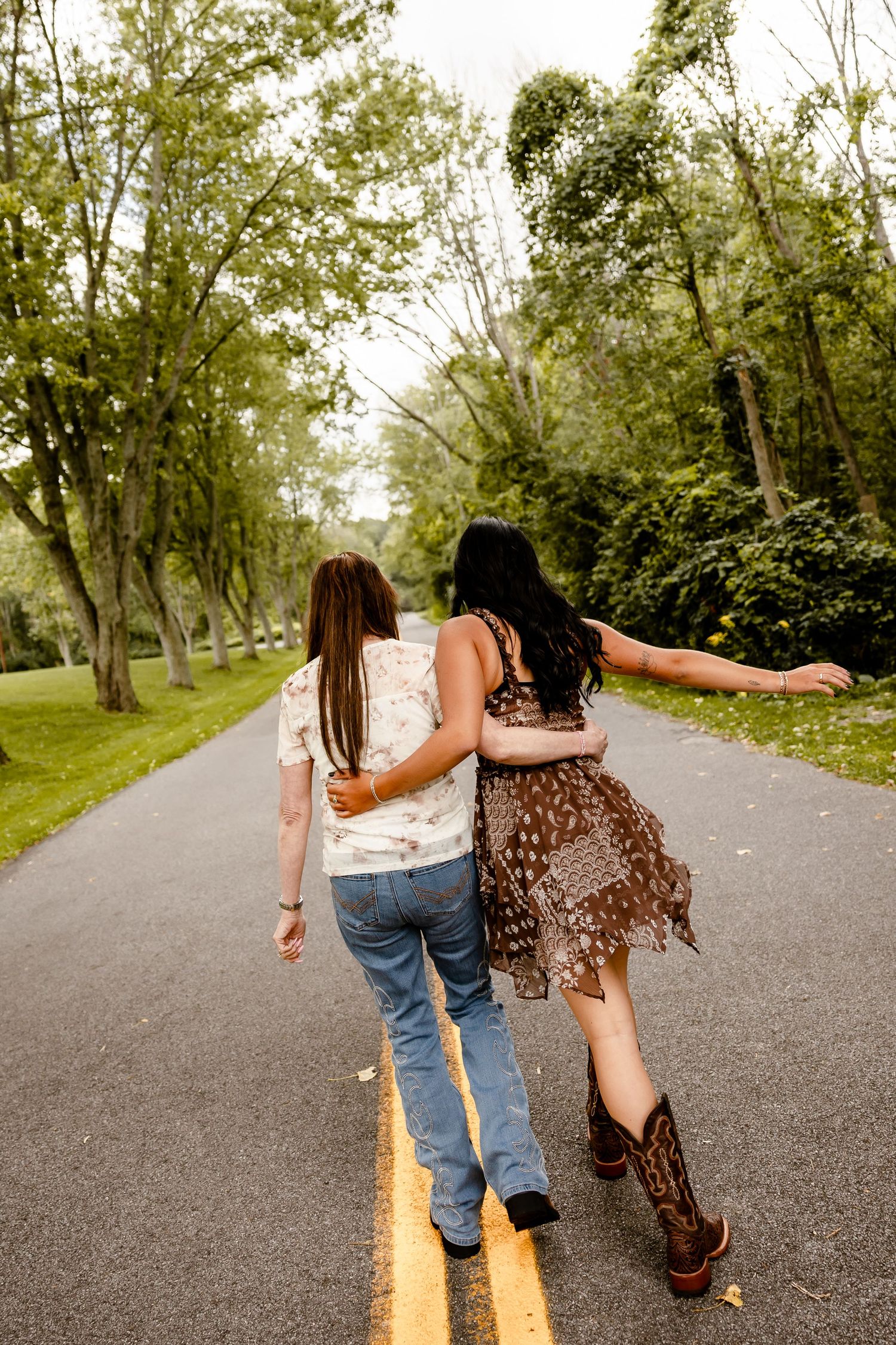 Two women walking arm in arm down a tree-lined road, one in jeans and one in a brown dress with cowboy boots.