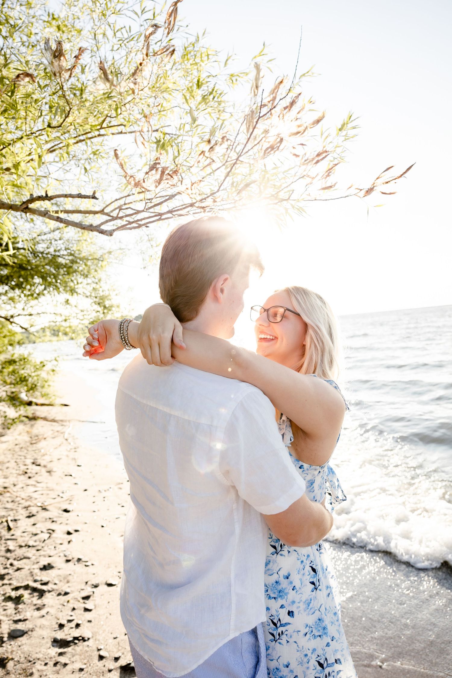 Couple embracing and smiling on a sunny beach with ocean waves and trees in the background.