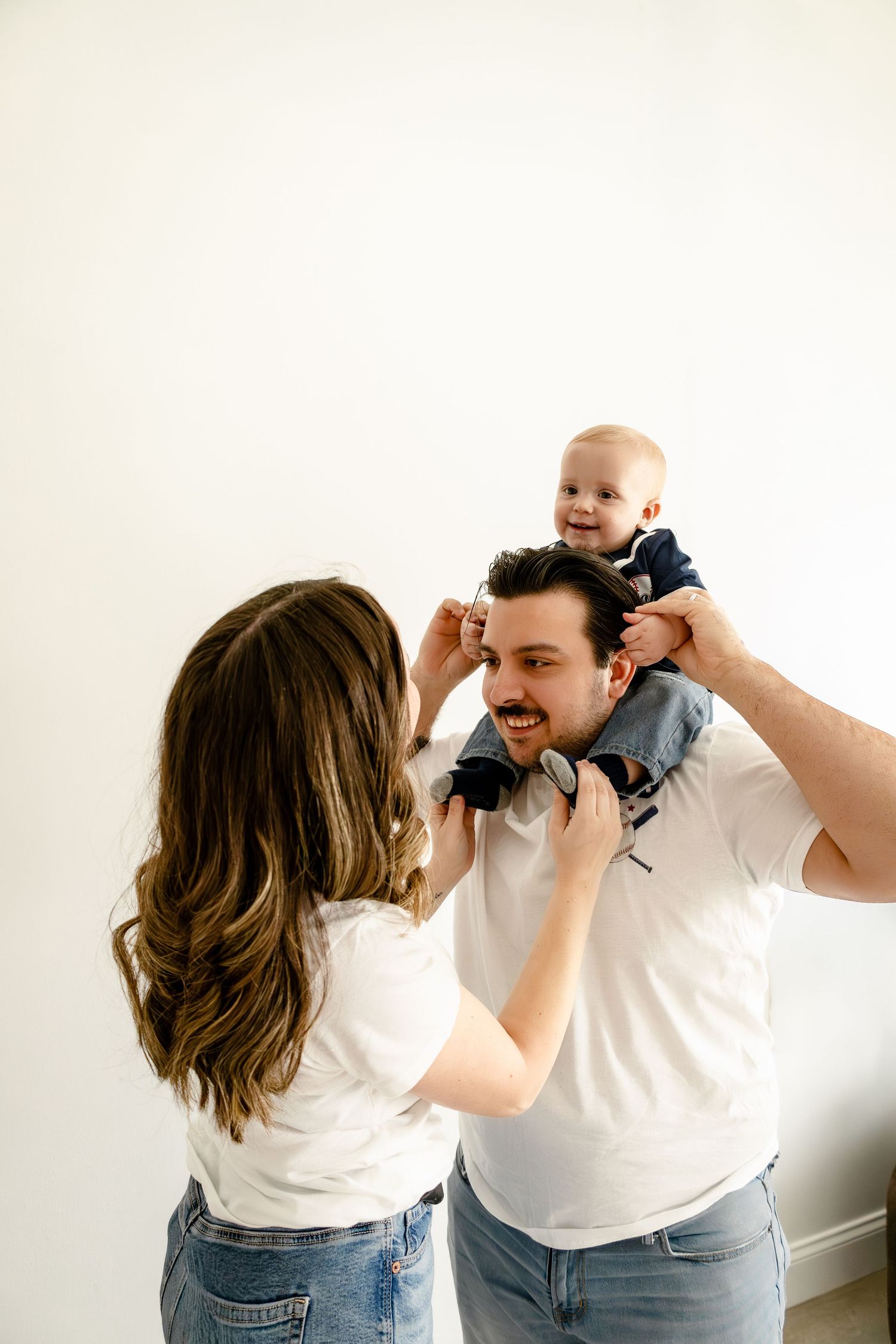 Happy young family with mother, father, and smiling baby in white tops and jeans against white wall.