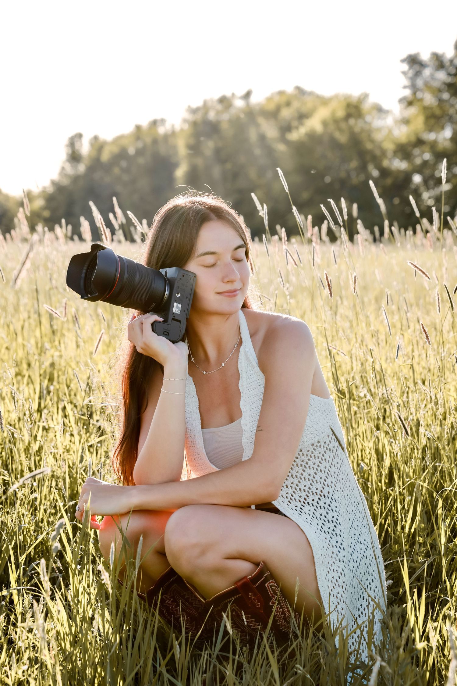 Young woman sitting in golden field grass holding DSLR camera, wearing white dress in warm sunlight.