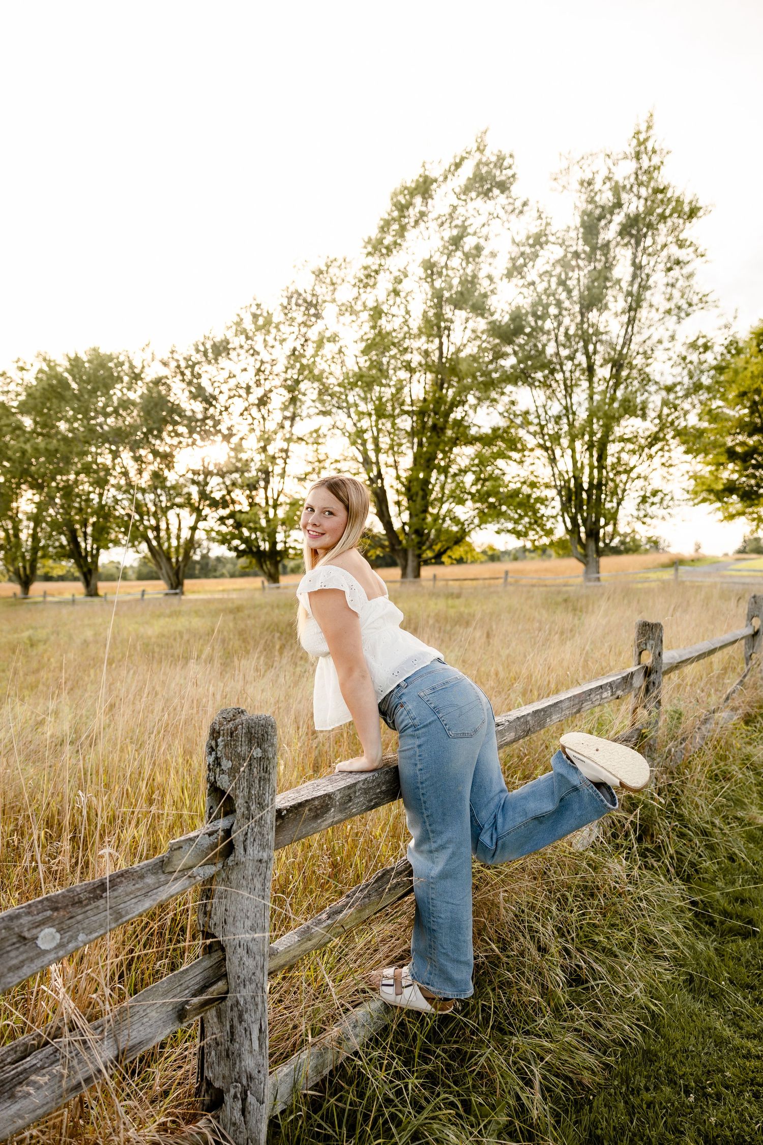 Young woman in white top and jeans leaning on rustic wooden fence in golden meadow with trees at sunset.