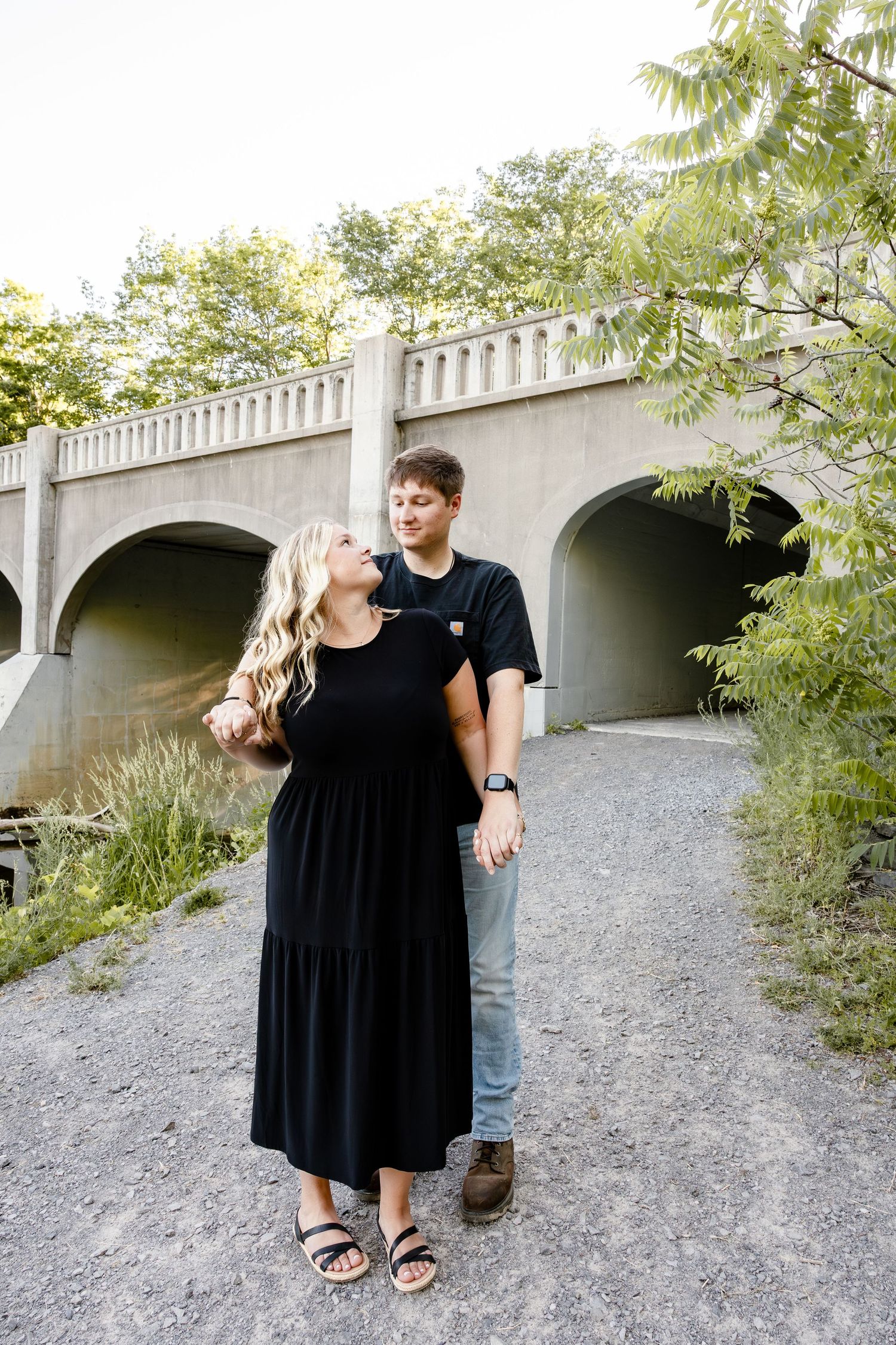 A couple in matching black outfits holds hands near a stone bridge surrounded by lush greenery on a gravel path.