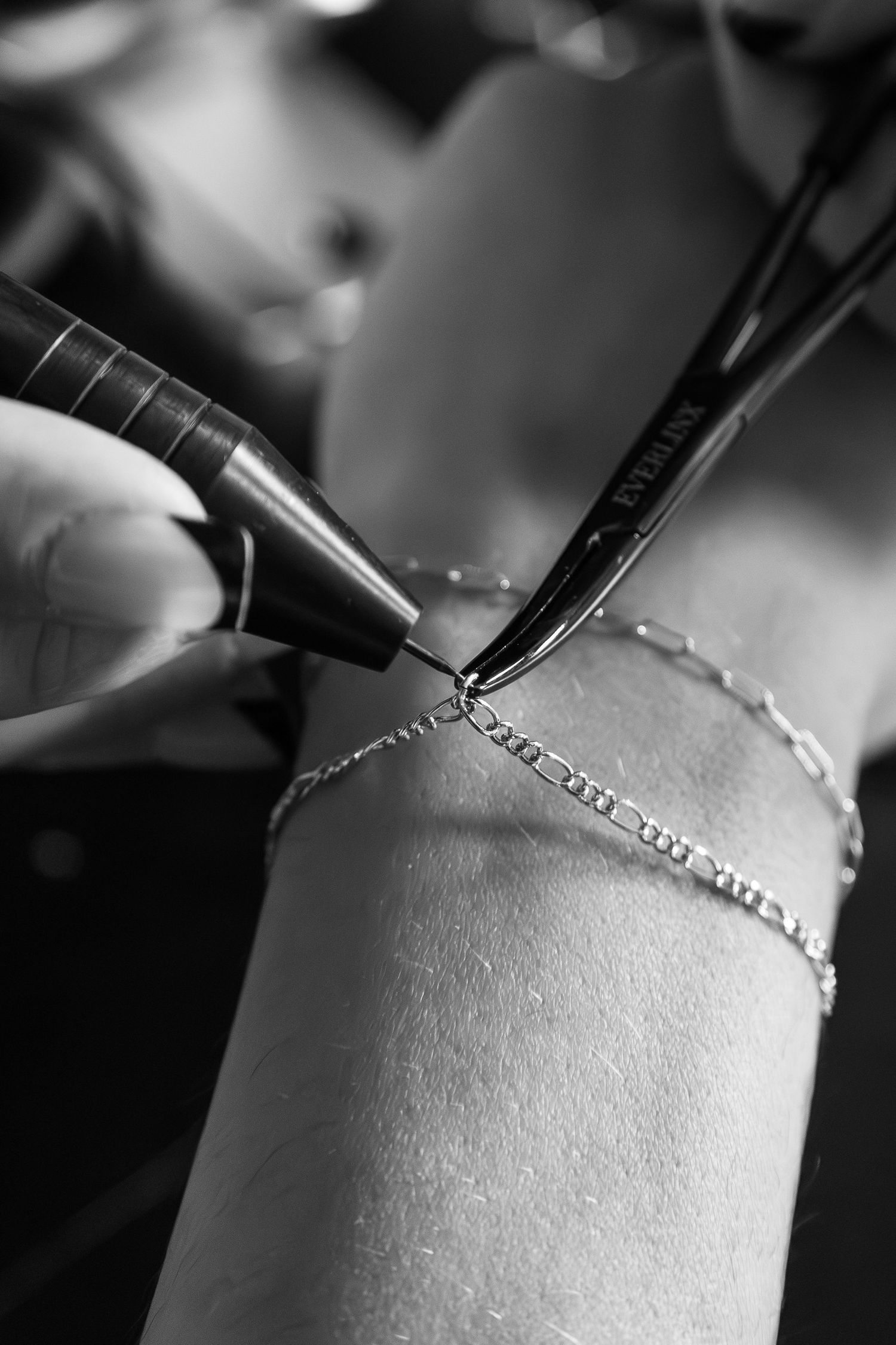 Black and white close-up of tattoo artist applying delicate wrist tattoo near silver chain bracelets.