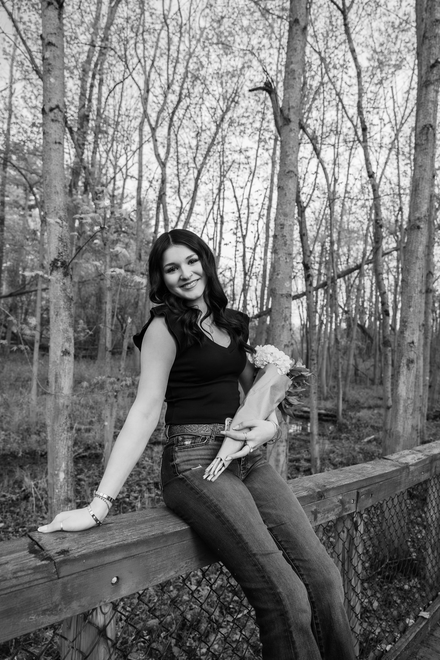 Black and white photo of a smiling young woman sitting on a wooden fence holding flowers in a forest setting.