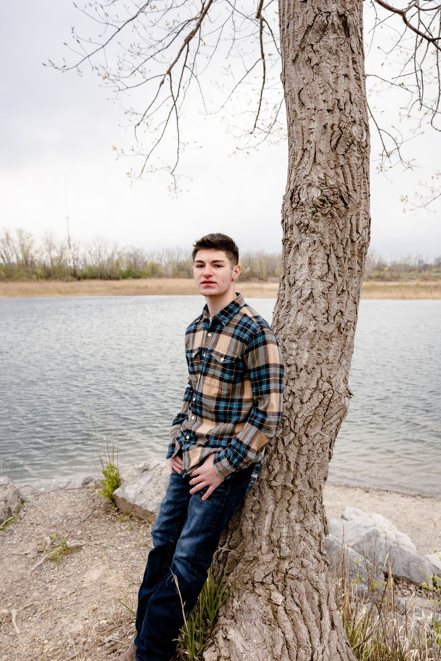 Young man in plaid shirt leaning against a tree by a lake on an overcast day.