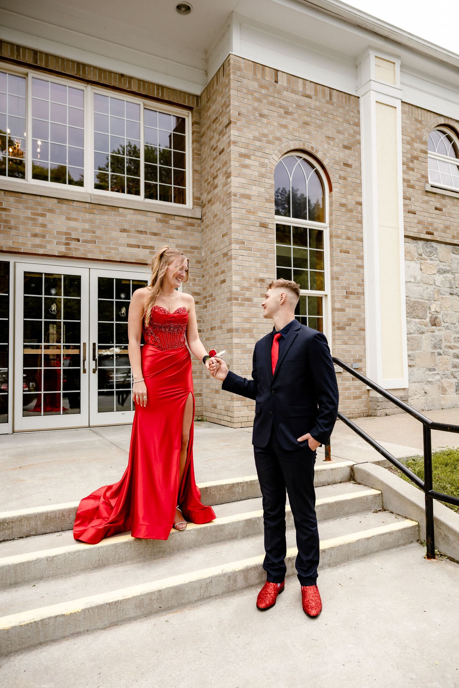 A couple in matching red and black prom attire pose on brick building steps, the woman in a red gown, man in a suit.