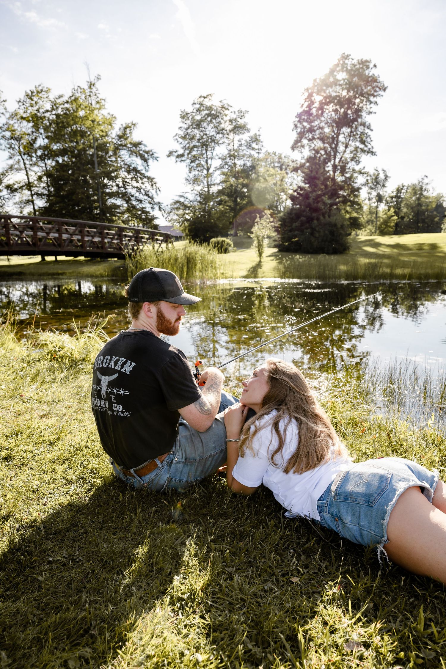 Couple relaxing by a pond in a sunny park, man sitting up while woman lies on grass, with a bridge in the background.