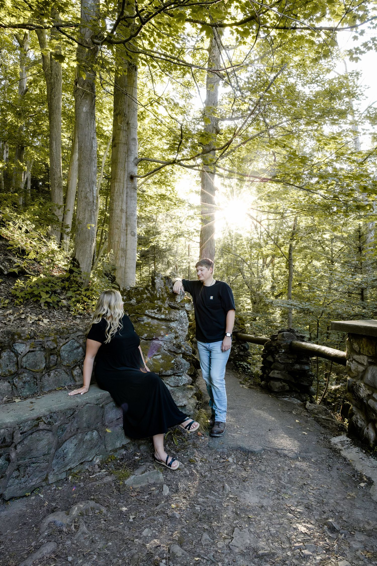 Couple in black outfits enjoying a sunlit forest path, woman seated on stone wall, man standing nearby.