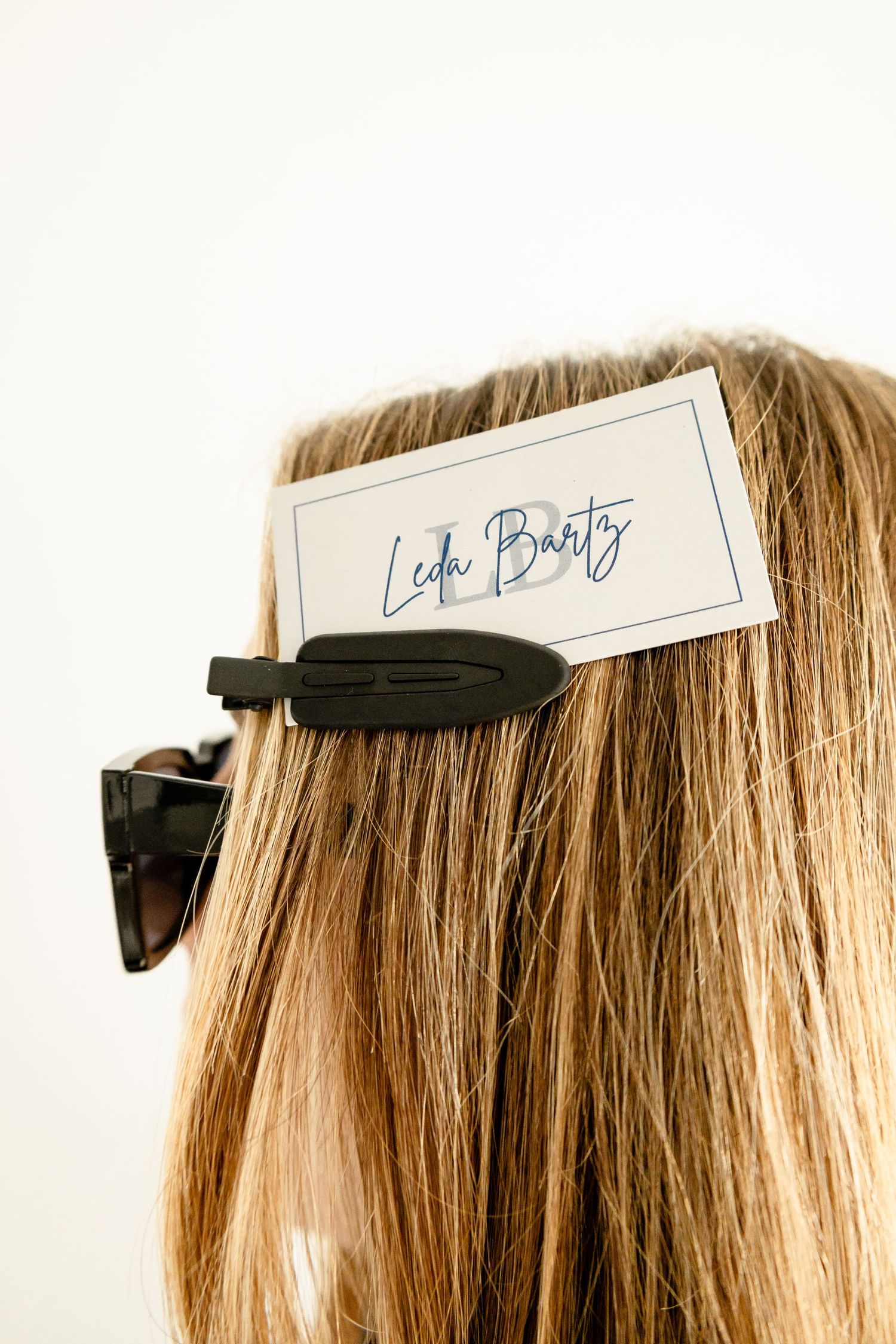 Woman with blonde hair wearing a black hair clip holding a name tag reading Leda Bartz, with sunglasses tucked in hair.
