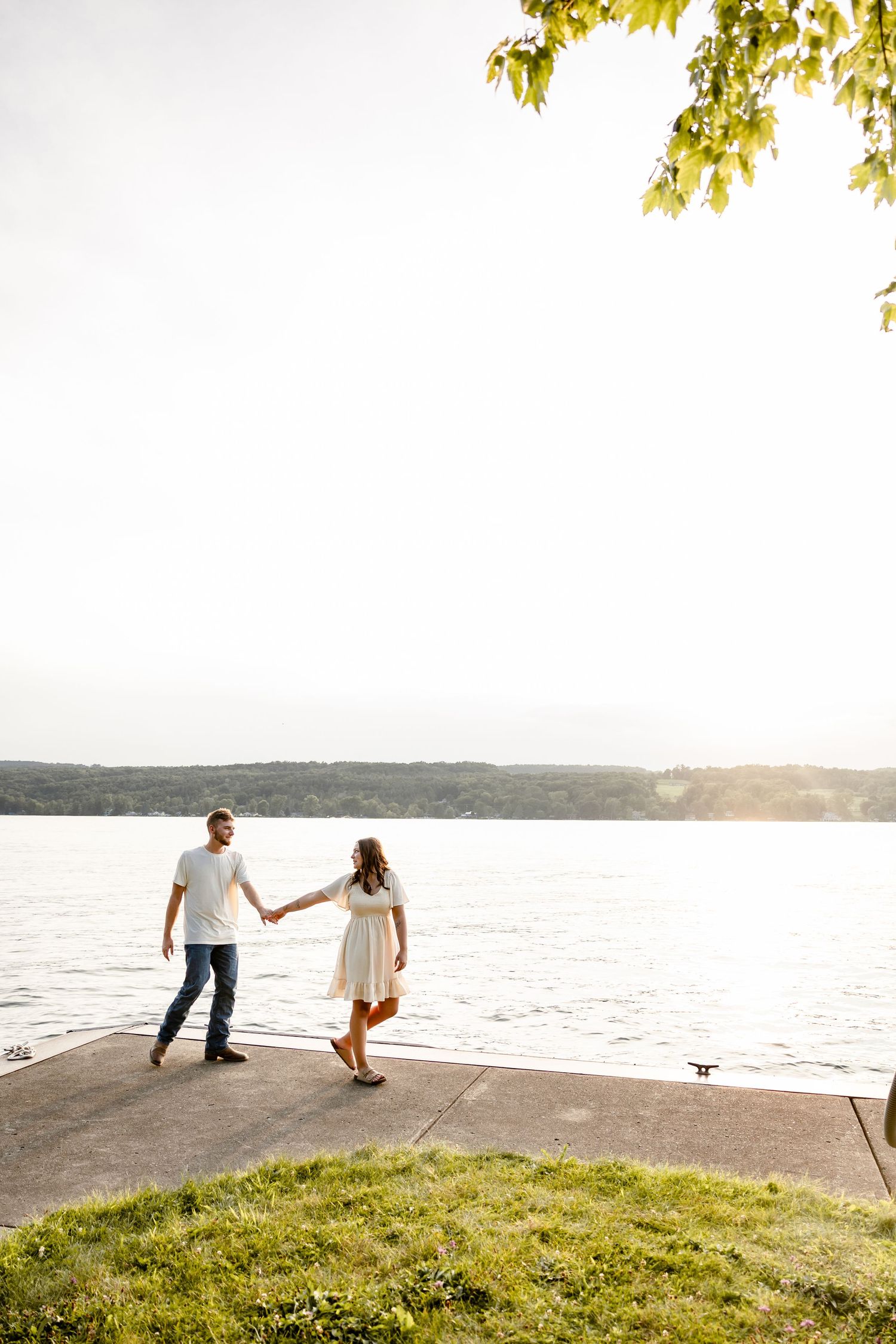 A couple holds hands and walks along a waterfront path at golden hour, surrounded by lush greenery and a serene lake view.
