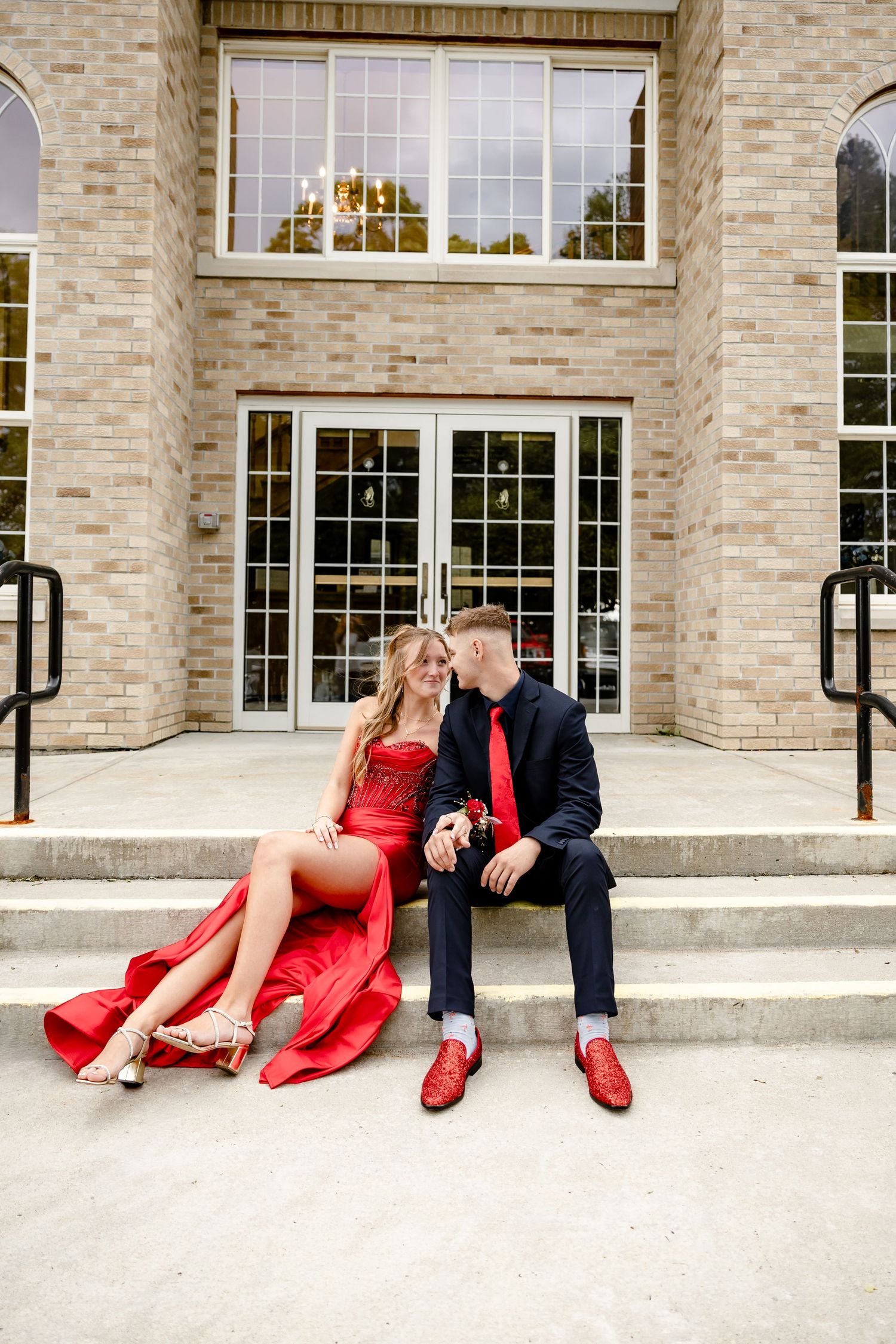 A couple in matching red outfits sit on building steps, sharing a romantic moment at what appears to be a prom event.