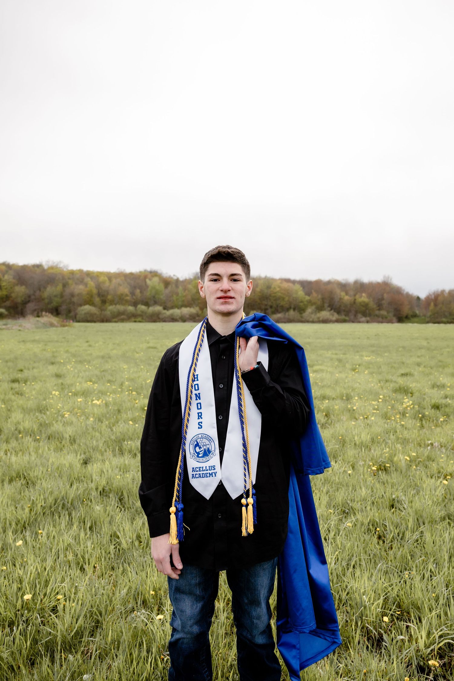 Young male graduate standing in green field wearing honors stole and holding blue graduation gown.