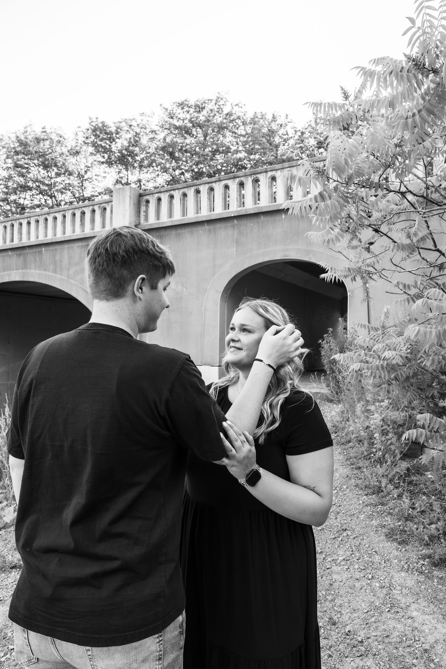 Black and white photo of a couple embracing near a stone bridge arch surrounded by trees.