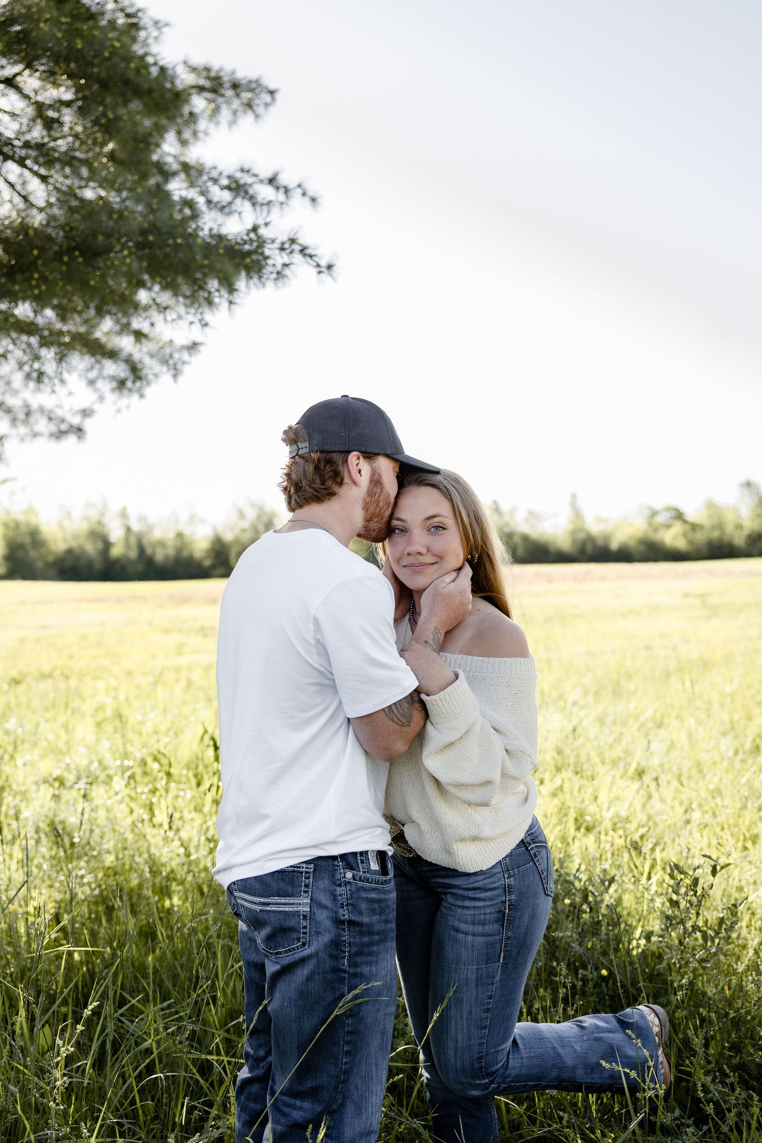 A couple embraces in a green field, man kissing woman's cheek as she smiles, both wearing jeans outdoors.