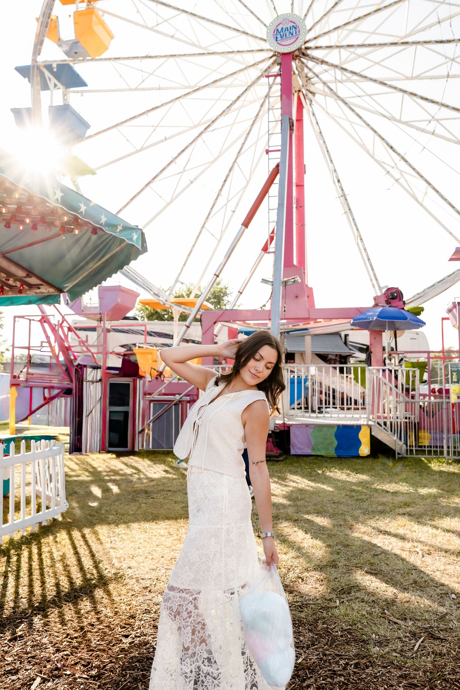 Woman in white lace dress posing at a colorful amusement park with a Ferris wheel in the background.