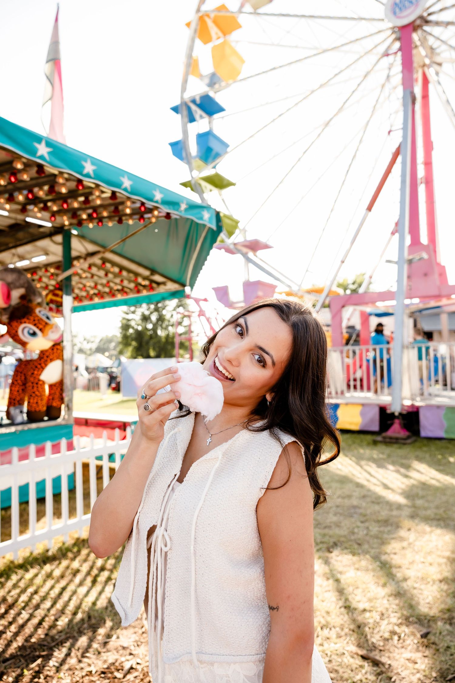 Young woman eating pink cotton candy at a colorful carnival with a Ferris wheel in the background.