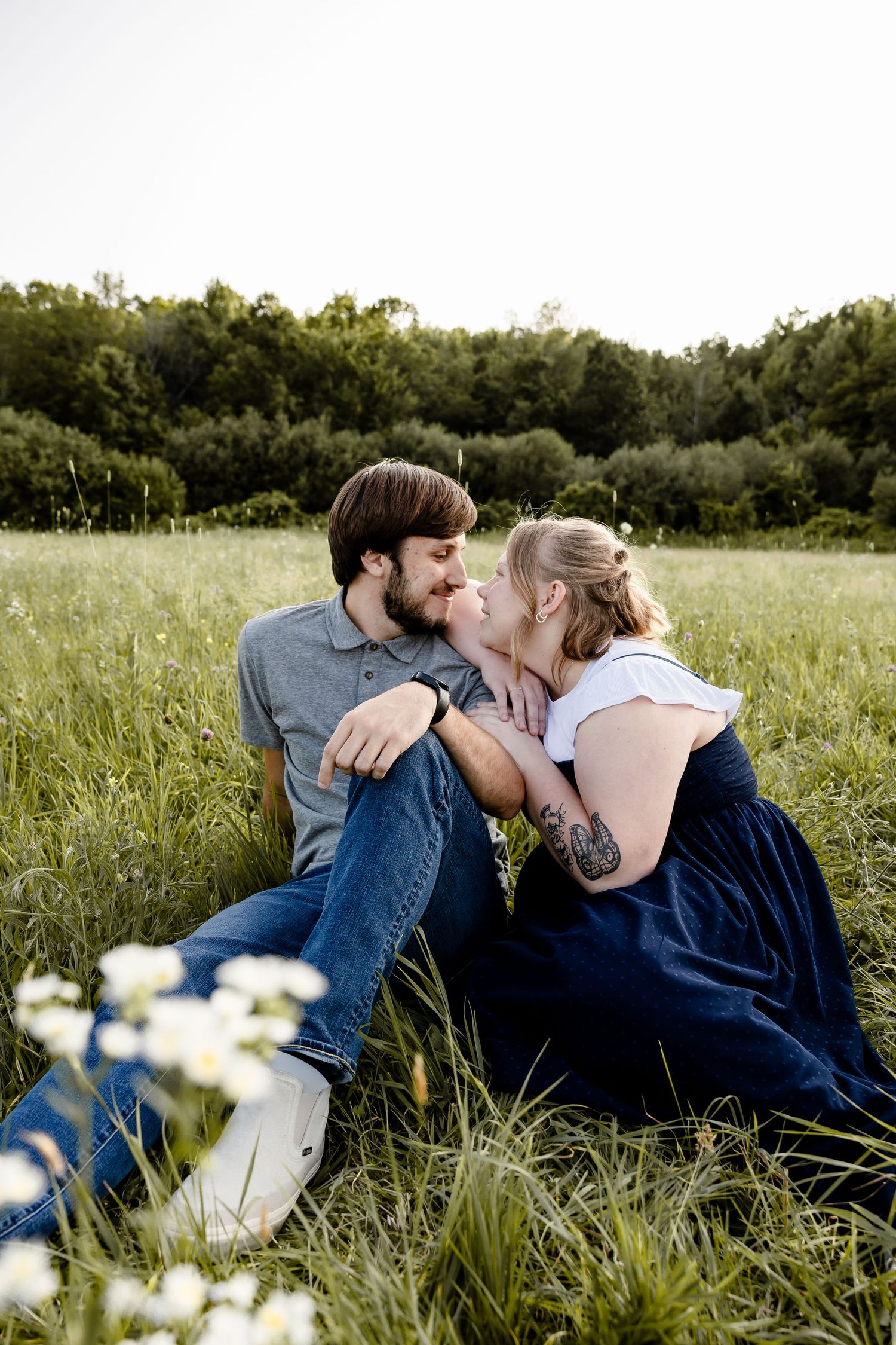 A couple shares a romantic moment sitting in a lush green field with wildflowers, nearly kissing during an outdoor engagement session.