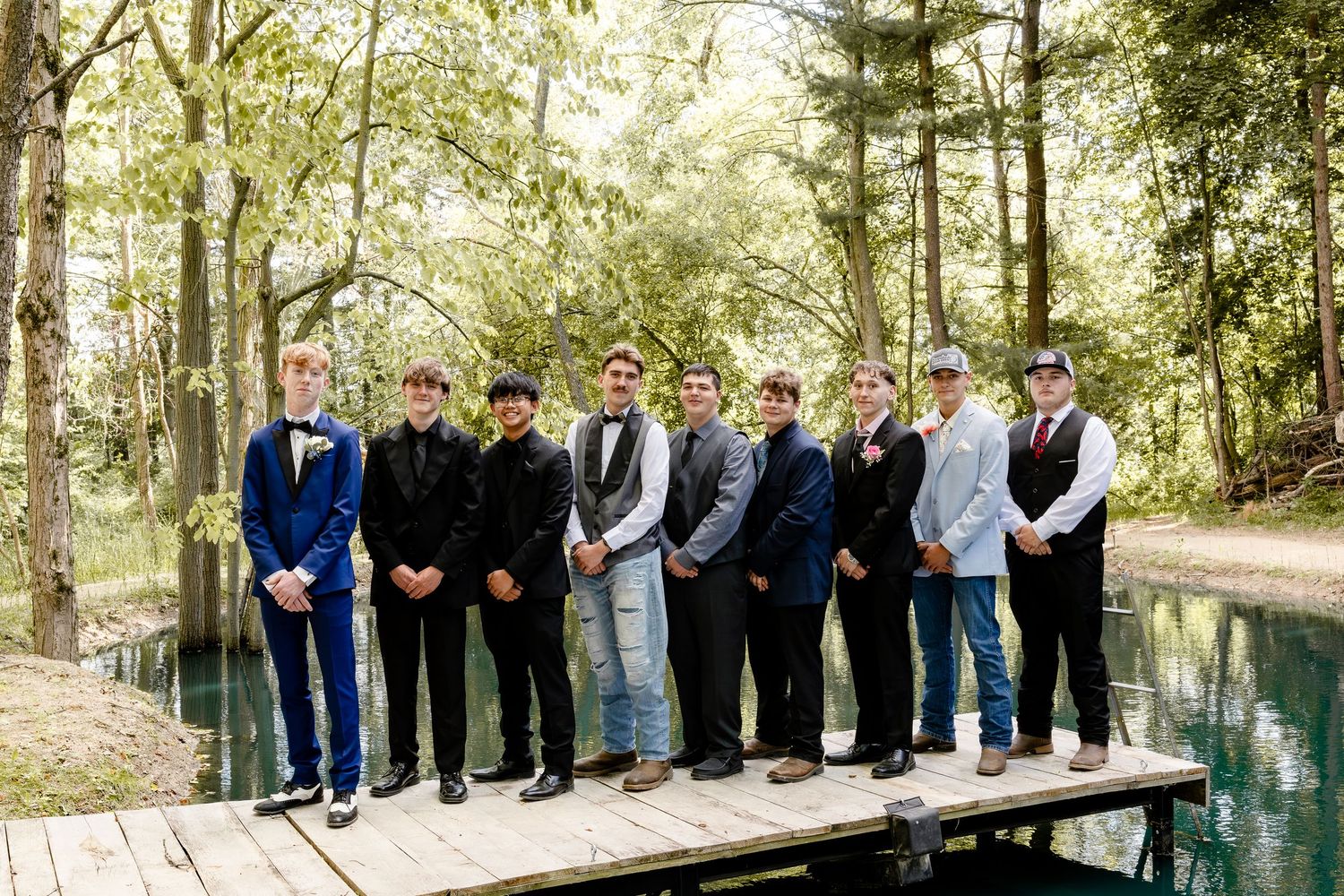 Group of teenage boys in formal attire posing on a wooden dock surrounded by lush green trees near a pond.