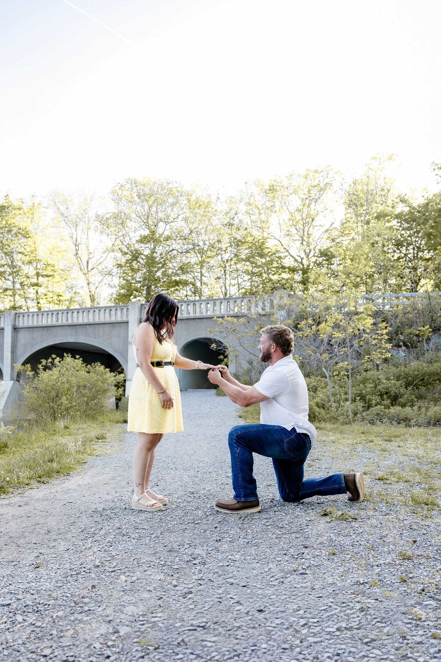 Man kneeling on gravel path proposing to woman in yellow dress near stone bridge outdoors.