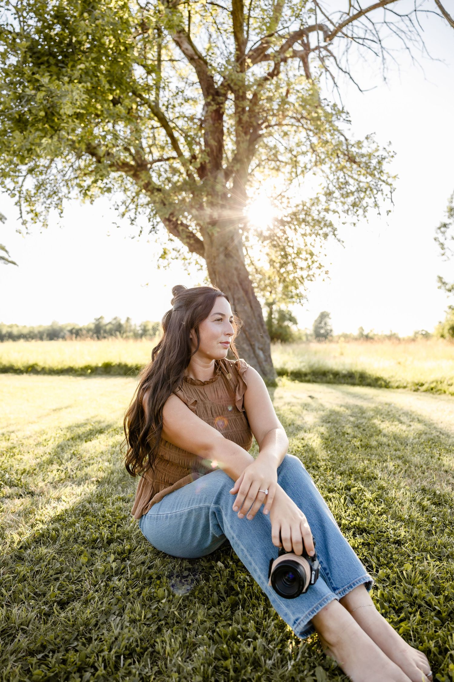 Woman sitting on grass under a sunlit tree, holding a camera, wearing a brown top and jeans in a serene outdoor setting.