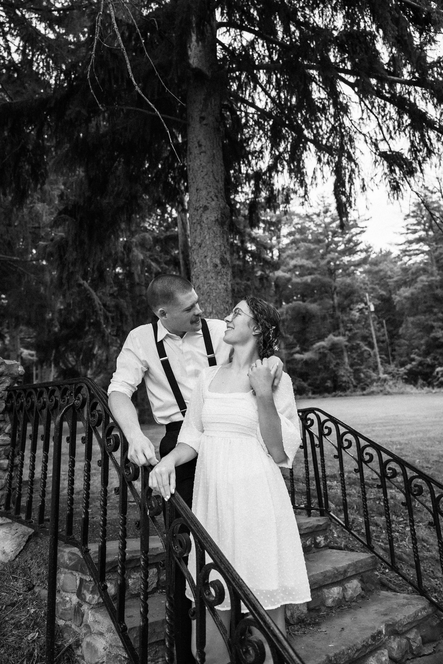 Black and white photo of a couple sharing a romantic moment on ornate iron stairs beneath a large pine tree.